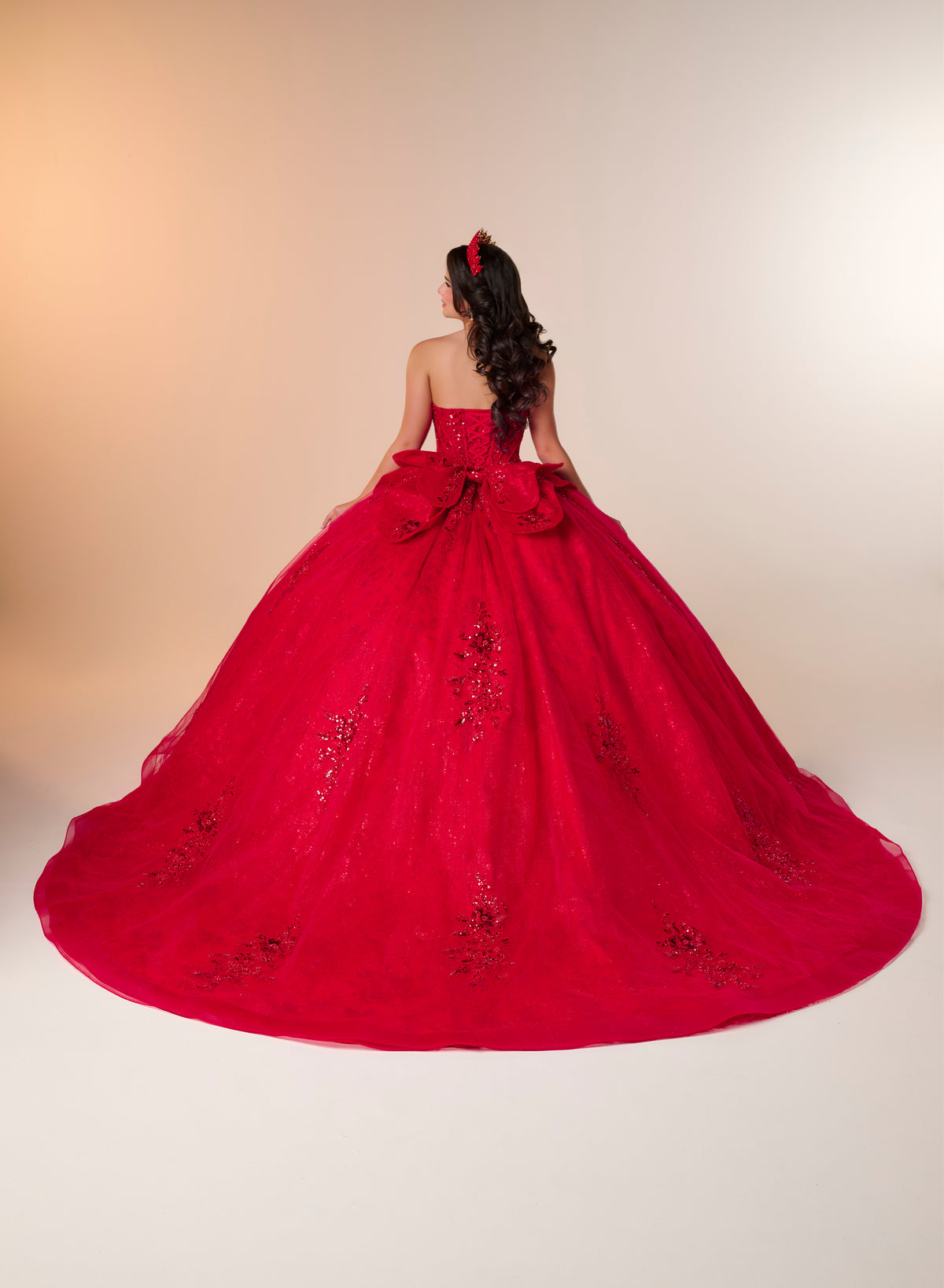A young woman viewed from behind wearing a voluminous bright red strapless ball gown with floral lace embroidery and a large bow at the back, standing against a warm beige studio backdrop. / A strapless ball gown featuring a beaded corset bodice, a voluminous glitter-tulle skirt with sequin appliques, and a textured ruffle accent on the back.