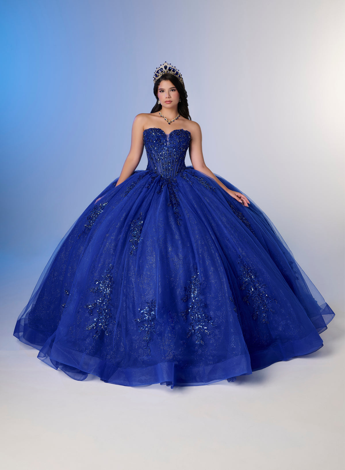 A young woman wearing a strapless royal blue quinceañera ball gown with a full tulle skirt and embroidered details, standing against a blue-to-white studio backdrop. / A strapless ball gown featuring a beaded corset bodice, a voluminous glitter-tulle skirt with sequin appliques, and a textured ruffle accent on the back.
