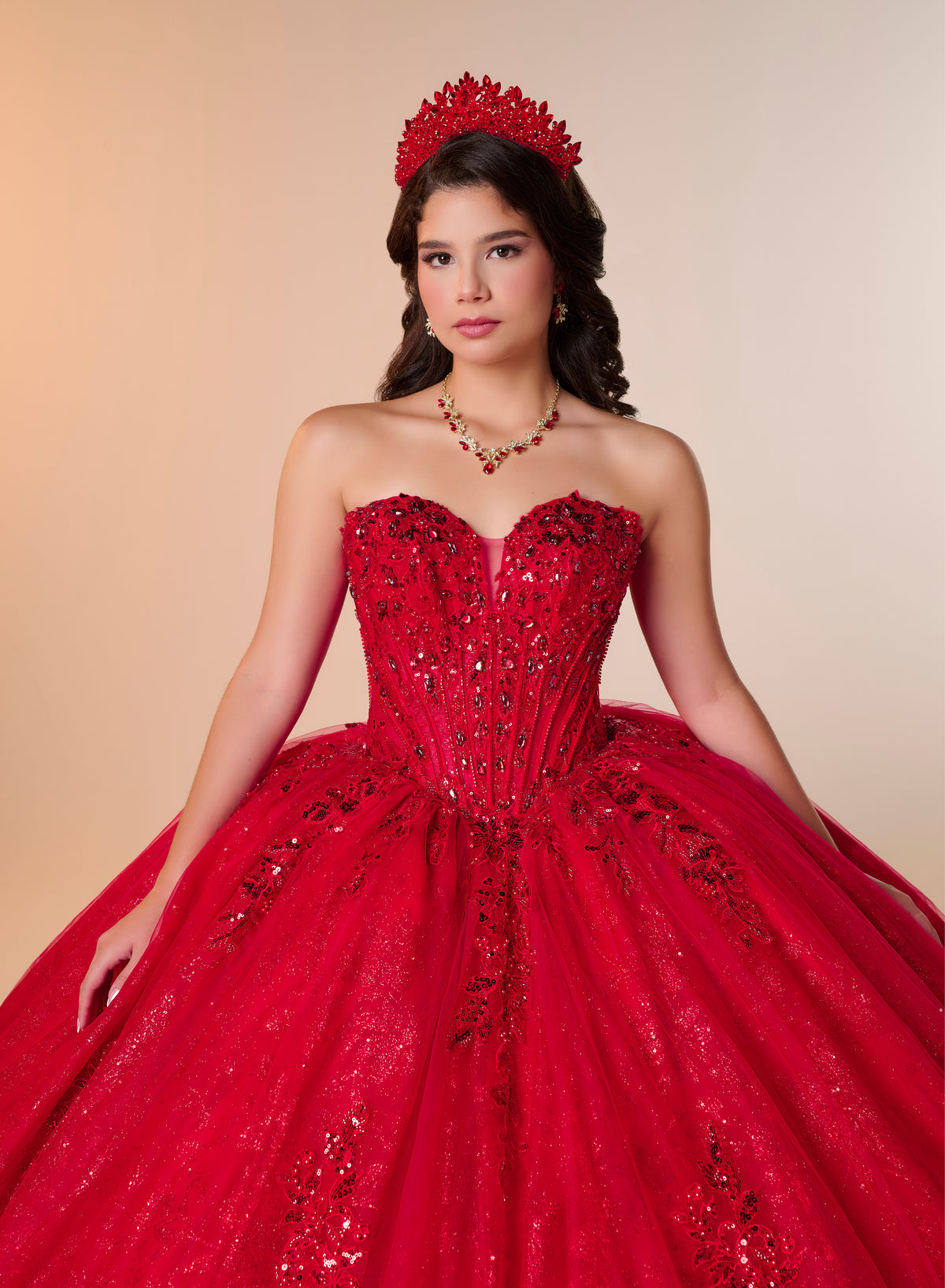 A young woman modeling a bright red strapless ball gown with a sweetheart neckline, heavily embellished with sequins and beadwork, paired with a matching red tiara and necklace against a warm beige studio background. / A strapless ball gown featuring a beaded corset bodice, a voluminous glitter-tulle skirt with sequin appliques, and a textured ruffle accent on the back.