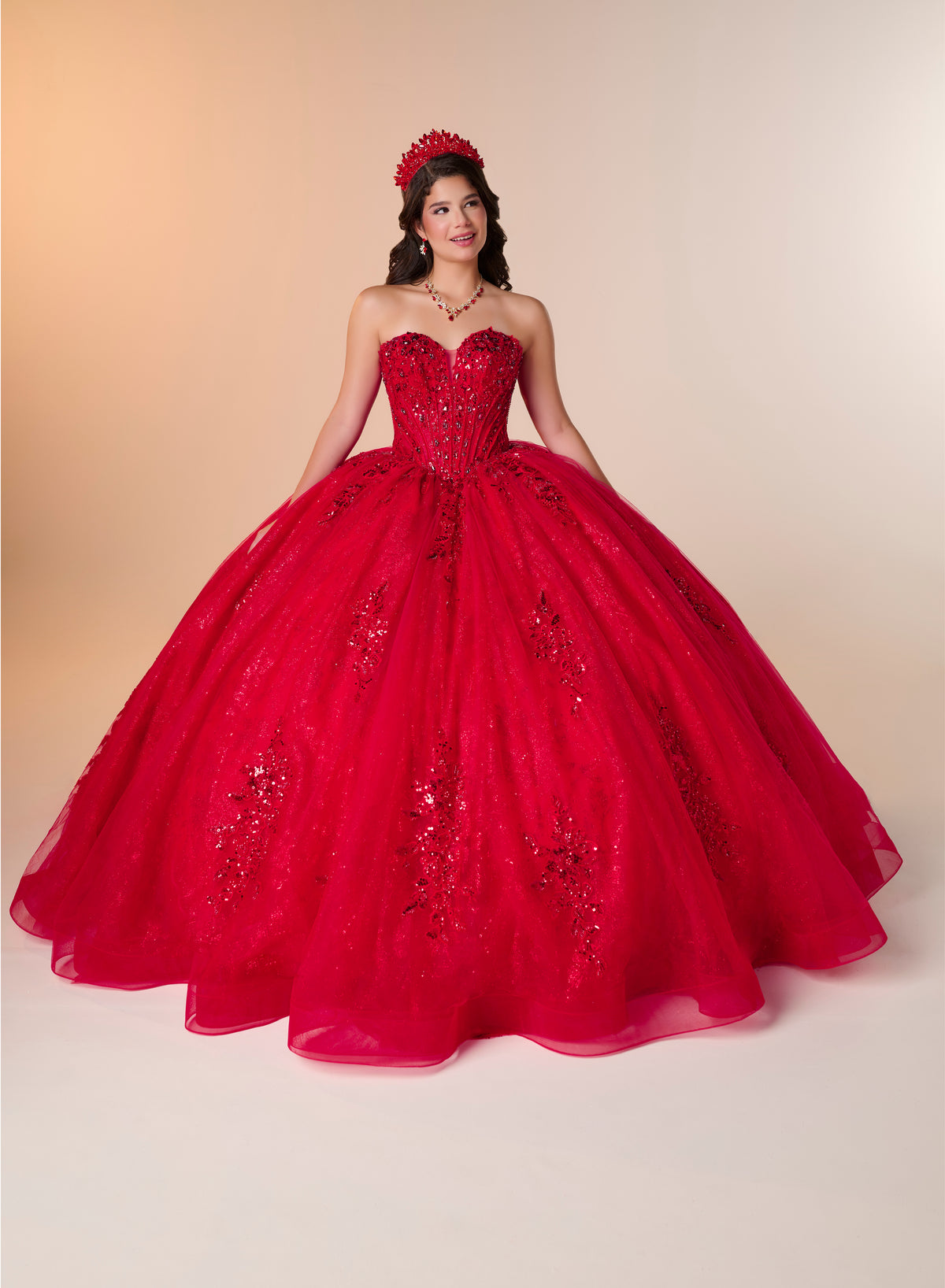 A young woman modeling a bright red strapless ball gown quinceañera dress with a sweetheart neckline, beaded corset bodice, and a voluminous tulle skirt, set against a warm beige studio background. / A strapless ball gown featuring a beaded corset bodice, a voluminous glitter-tulle skirt with sequin appliques, and a textured ruffle accent on the back.