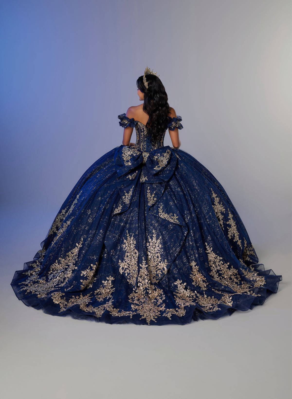A woman photographed from behind wearing an elaborate royal blue ball gown with extensive gold floral embroidery, set against a neutral studio background. / An off-the-shoulder ball gown with ruffled cap sleeves, a heavily embroidered metallic bodice, and a voluminous glitter-tulle skirt adorned with intricate baroque-style appliques and a matching oversized back bow.