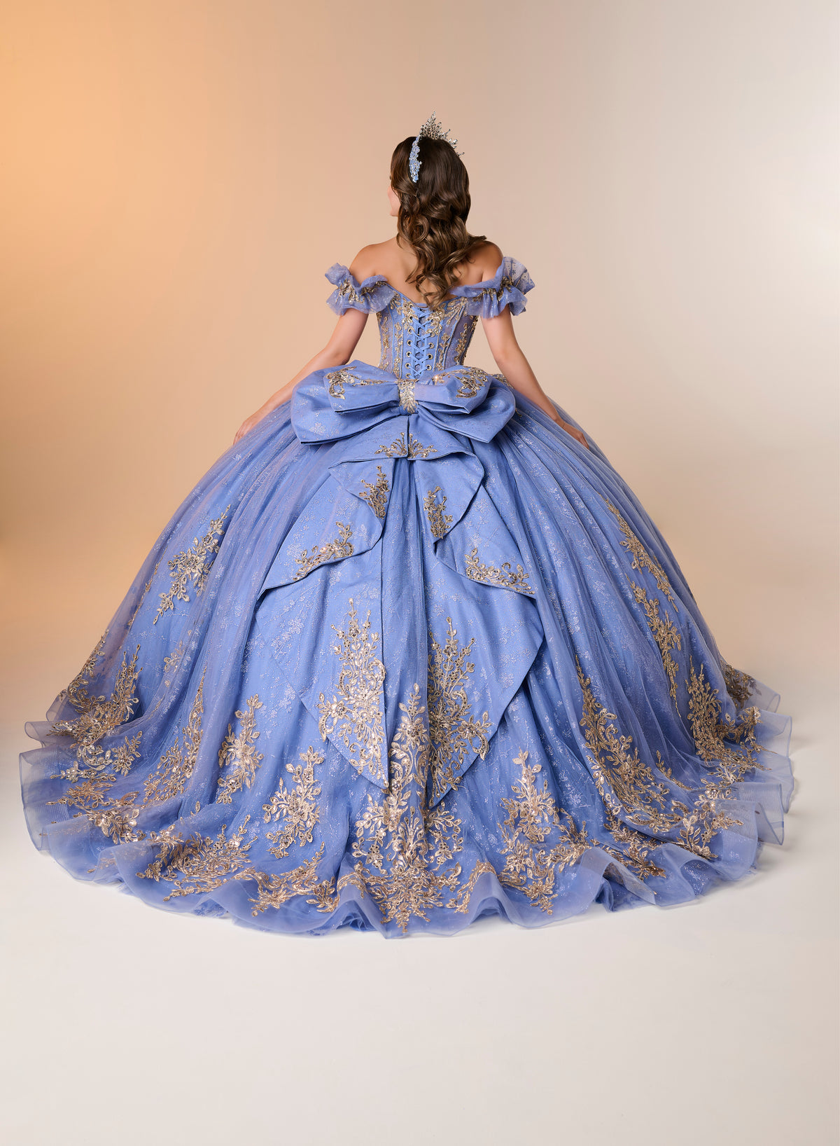 A woman viewed from behind wearing an elaborate light blue ball gown with gold floral embroidery and a large bow at the back, photographed against a plain beige studio background. / An off-the-shoulder ball gown with ruffled cap sleeves, a heavily embroidered metallic bodice, and a voluminous glitter-tulle skirt adorned with intricate baroque-style appliques and a matching oversized back bow.