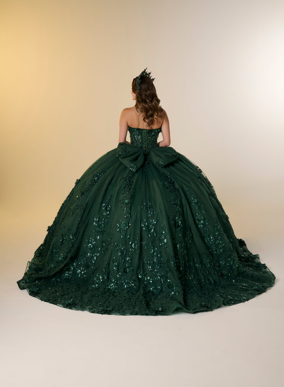 A model viewed from behind wearing a strapless dark green ball gown with a large bow at the back of the bodice and a voluminous tulle skirt covered in floral lace and sequin embellishments, set against a warm beige studio background. / A strapless sweetheart ball gown featuring a fully beaded corset bodice, a lace-up back, and a voluminous tulle skirt layered with shimmering sequin floral details and an oversized back bow, paired with a matching beaded choker necklace.