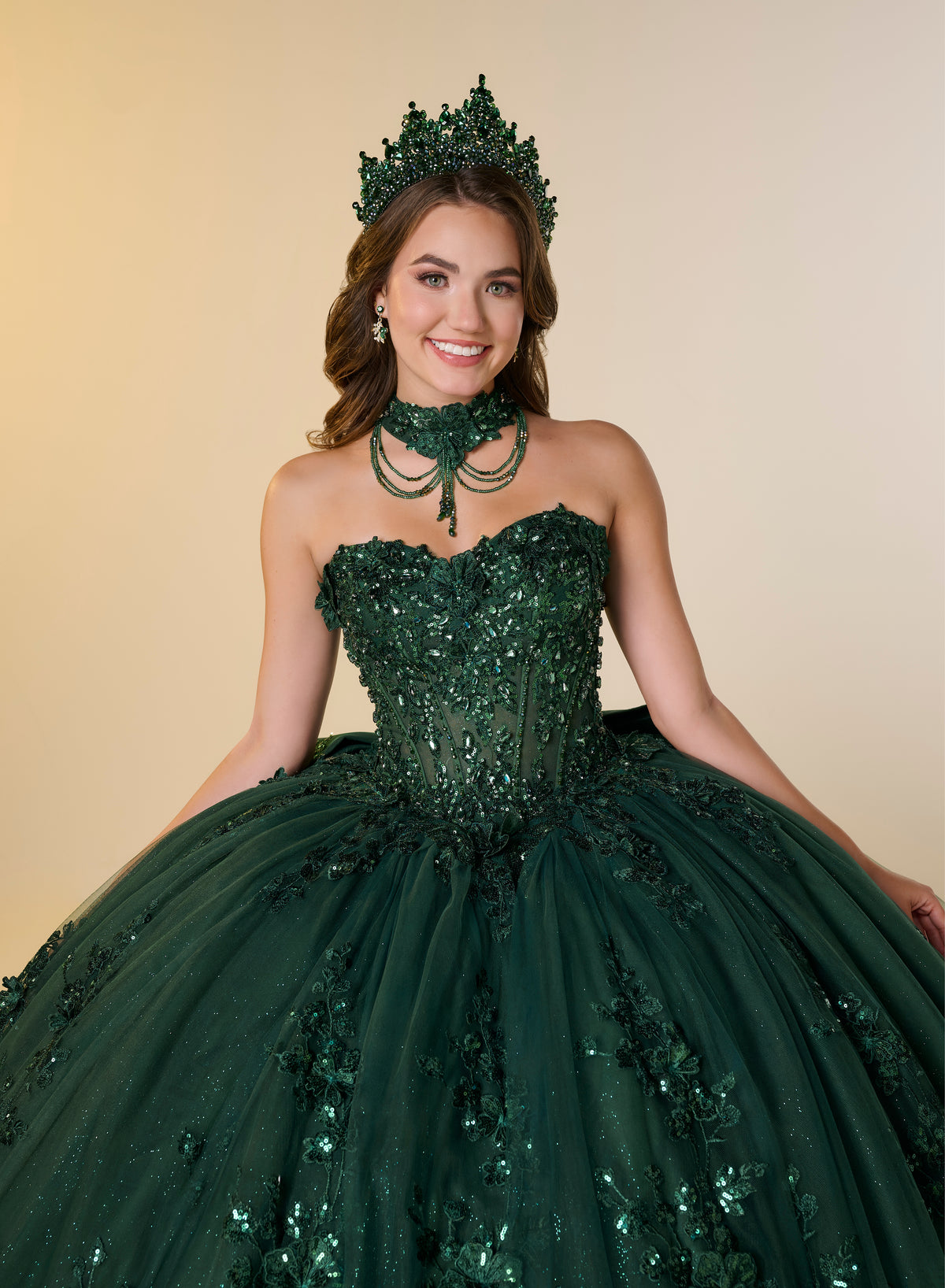 A woman wearing a voluminous dark green quinceañera ball gown with sequined floral appliqué, a matching green choker necklace, and a green tiara, posed against a warm beige studio background. / A strapless sweetheart ball gown featuring a fully beaded corset bodice, a lace-up back, and a voluminous tulle skirt layered with shimmering sequin floral details and an oversized back bow, paired with a matching beaded choker necklace.