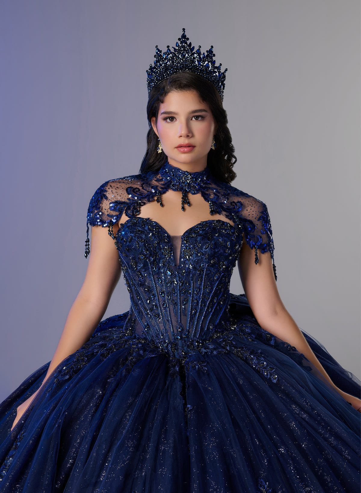 A woman wearing an elaborate dark blue ball gown with a sheer lace high neckline, beaded corset bodice, and voluminous tulle skirt, styled with a matching jeweled crown against a plain gray studio background. / Ball gown with a lace-detailed back, corset lacing, and a large bow at the waist. The full skirt has a subtle sparkle and elegant embroidery.