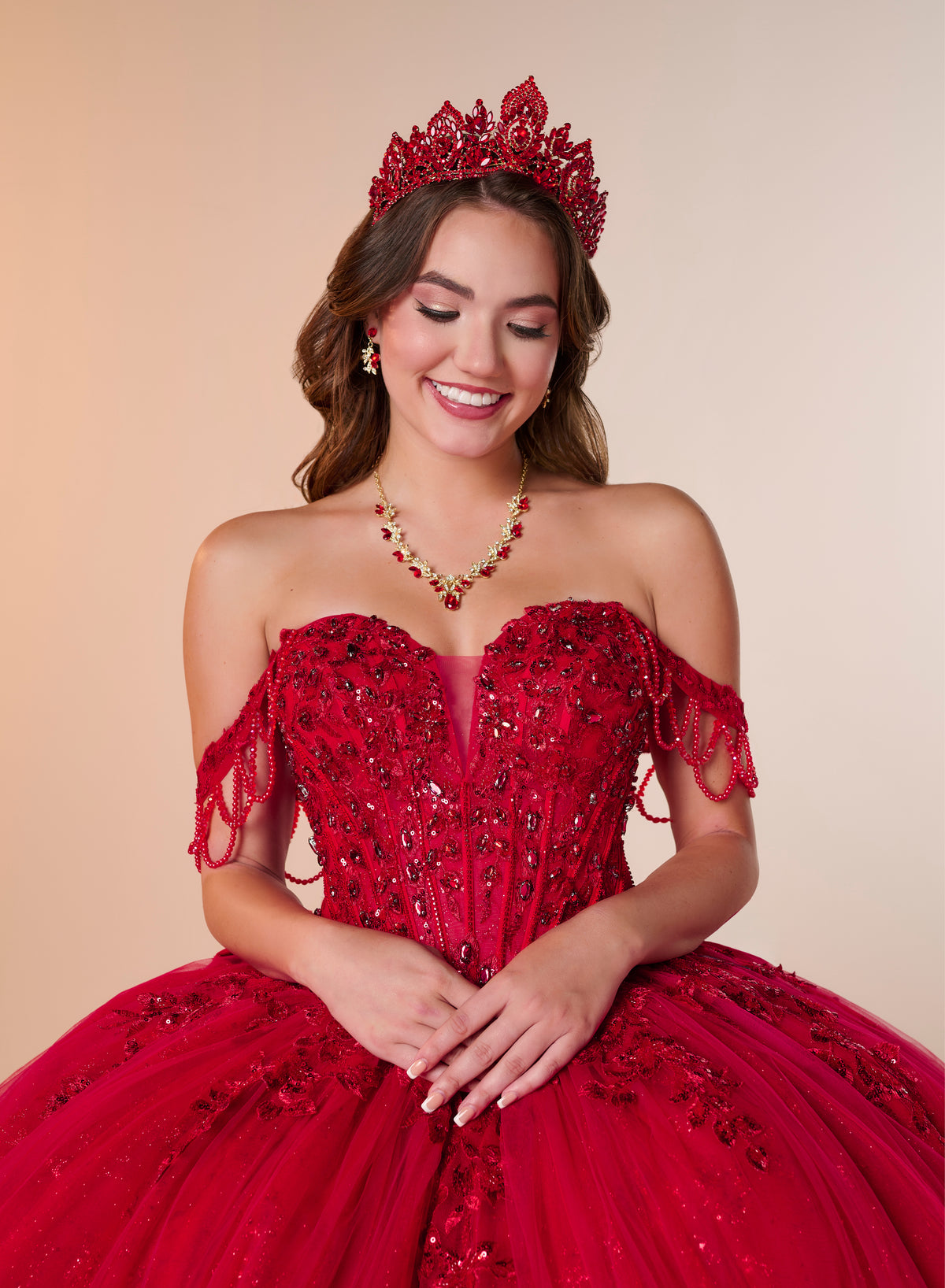 A smiling young woman wearing a bright red off-the-shoulder quinceañera dress with heavy beadwork, a matching red tiara, and a red beaded necklace, posed against a warm beige studio background. / Ball gown with a lace-detailed back, corset lacing, and a large bow at the waist. The full skirt has a subtle sparkle and elegant embroidery.