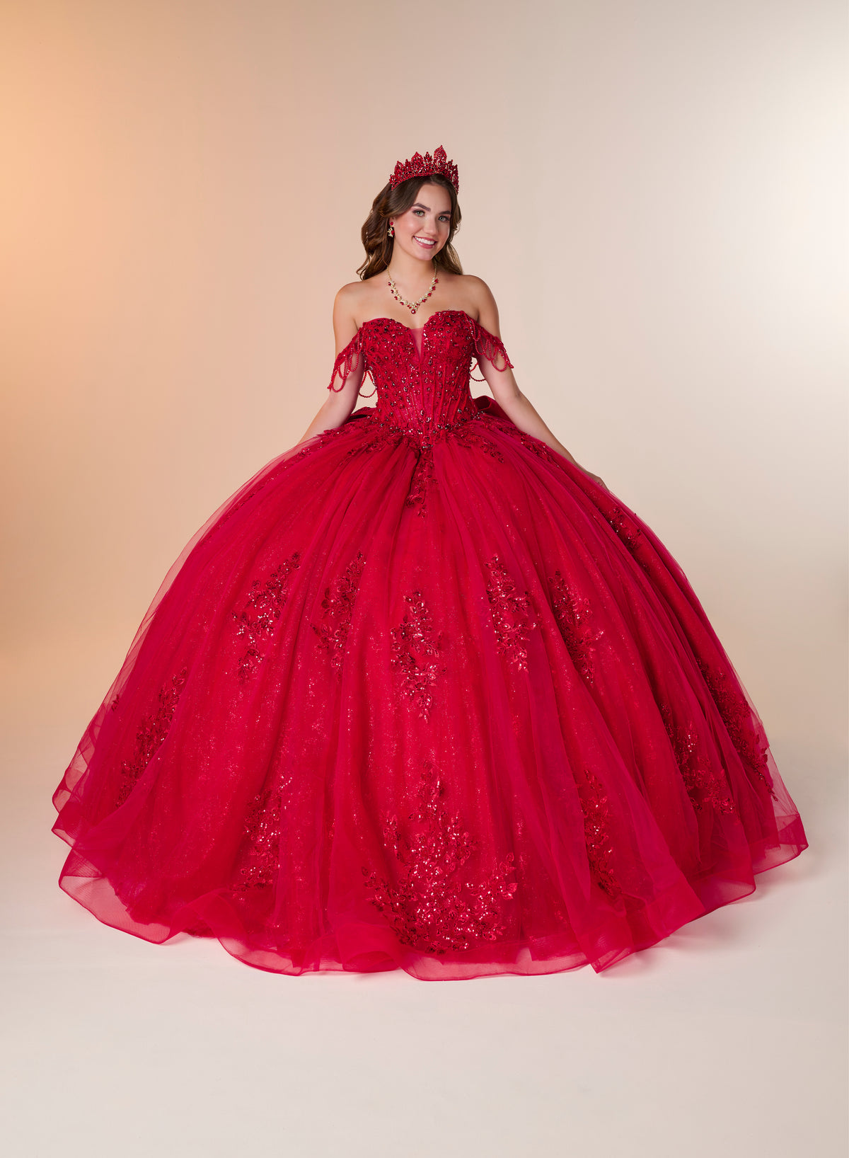 A young woman wearing a voluminous off-the-shoulder red quinceañera ball gown with floral lace applique and a matching red tiara, posed against a warm beige studio backdrop. / Ball gown with a lace-detailed back, corset lacing, and a large bow at the waist. The full skirt has a subtle sparkle and elegant embroidery.