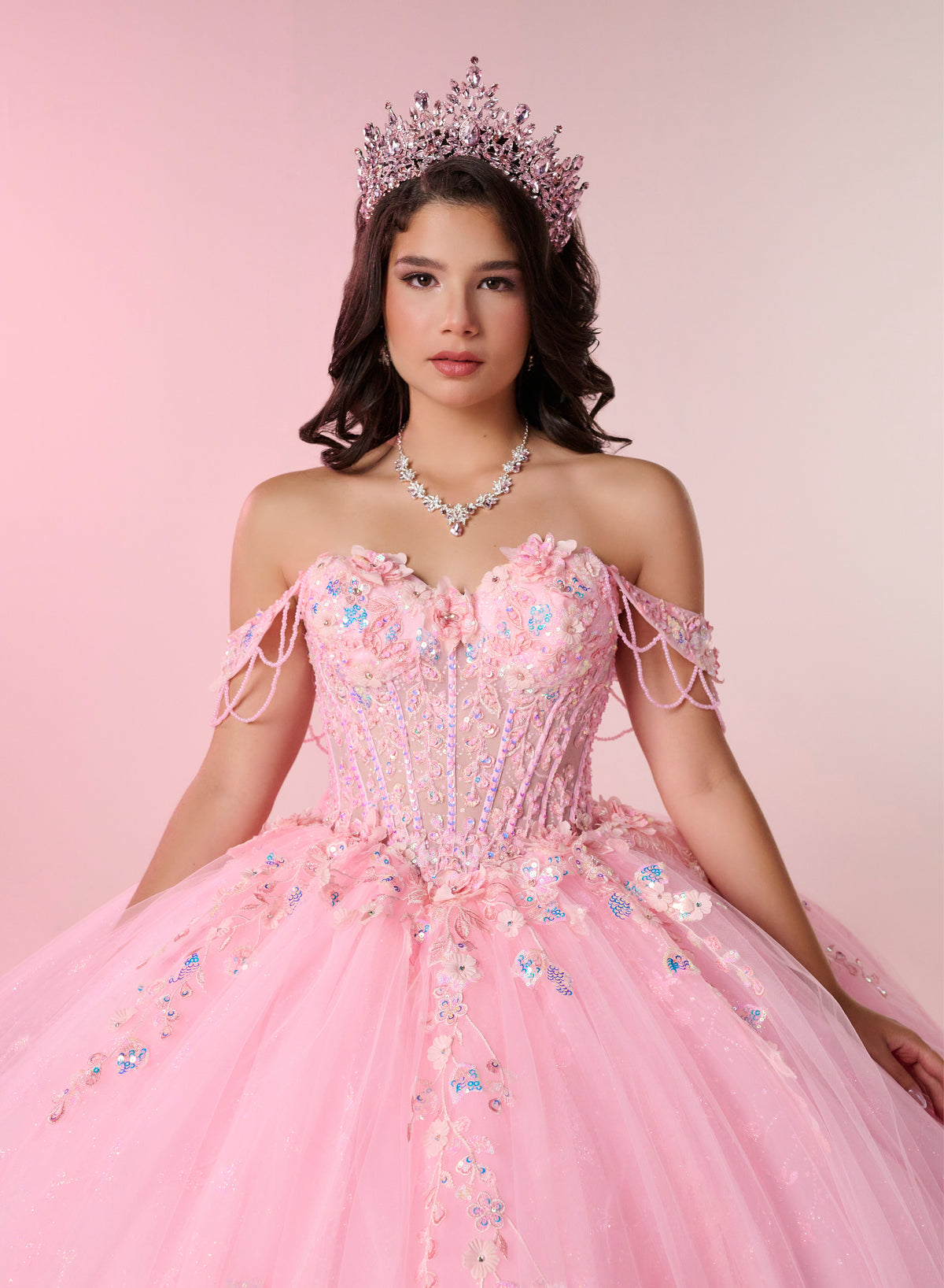 A woman wearing a pink off-the-shoulder quinceañera dress with floral embroidery, a matching tiara, and a pearl necklace, posed against a solid pink studio background. / An off-shoulder ball gown with a lace-up back, decorative bow, and floral embellishments on a full, layered skirt.