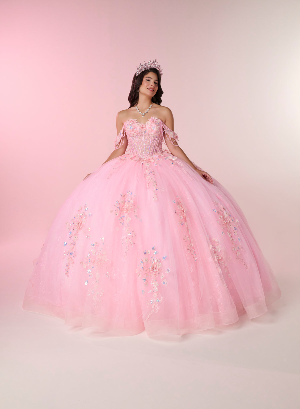 A young woman wearing a voluminous light pink ball gown with floral embroidery and a tiara, posing against a soft pink studio backdrop. / An off-shoulder ball gown with a lace-up back, decorative bow, and floral embellishments on a full, layered skirt.