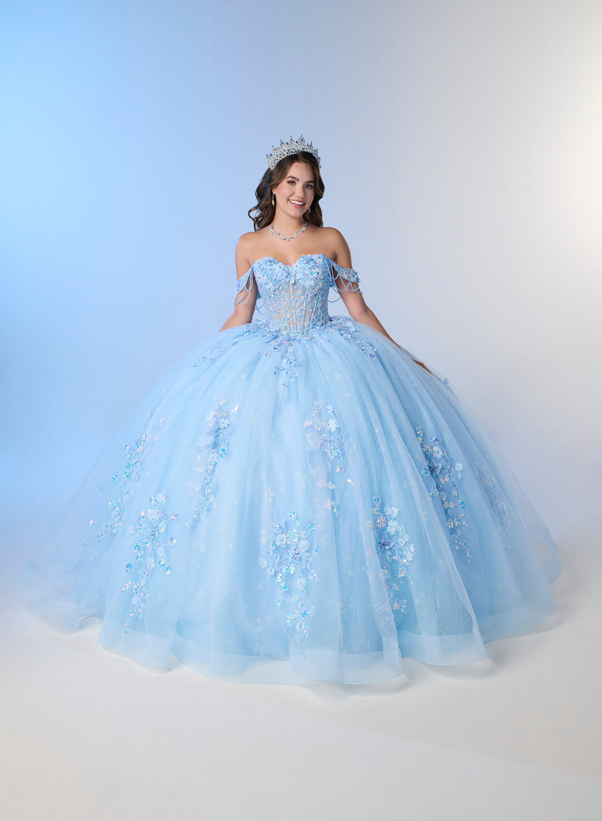A young woman wearing a voluminous light blue off-the-shoulder ball gown with floral embroidery and beadwork, standing against a soft blue-to-white studio backdrop. She is also wearing a small tiara. / An off-shoulder ball gown with a lace-up back, decorative bow, and floral embellishments on a full, layered skirt.