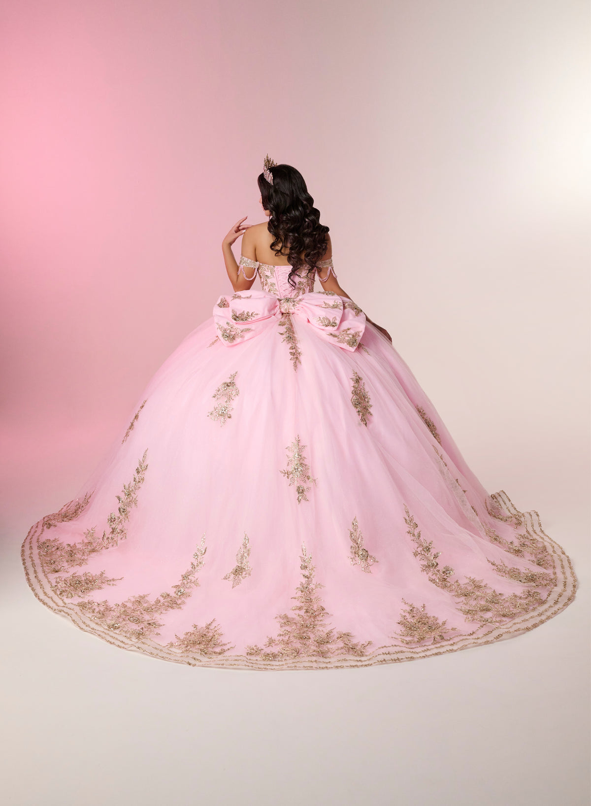 A woman viewed from behind wearing an elaborate light pink ball gown with gold floral embroidery and a very full train, posed against a soft pink studio background. / Two-piece off-shoulder dress with a fitted, embellished bodice and a short, form-fitting skirt, featuring a sweetheart neckline, sparkling embroidered details, detachable arm drapes, and a full tulle skirt with a sweeping train.