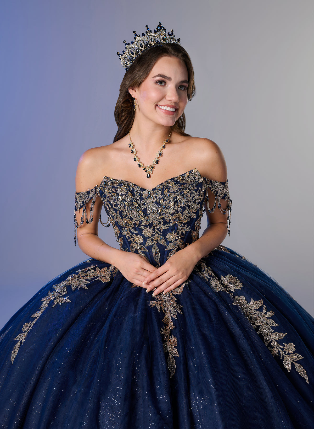 A young woman modeling an off-the-shoulder navy blue ball gown with intricate gold floral embroidery, wearing a matching tiara and necklace against a solid gray studio background. / Two-piece off-shoulder dress with a fitted, embellished bodice and a short, form-fitting skirt, featuring a sweetheart neckline, sparkling embroidered details, detachable arm drapes, and a full tulle skirt with a sweeping train.