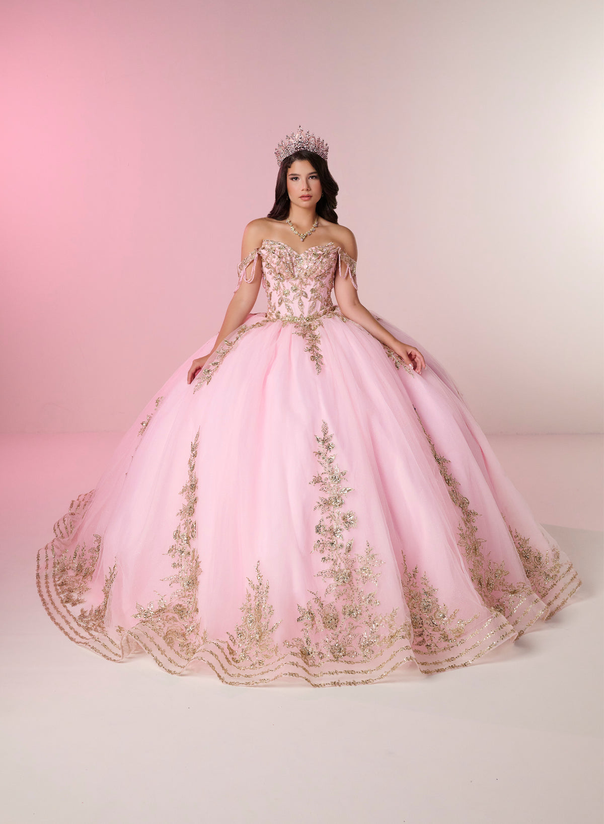 A woman wearing a voluminous light pink ball gown with gold floral embroidery and a matching tiara, posed against a soft pink studio backdrop. / Two-piece off-shoulder dress with a fitted, embellished bodice and a short, form-fitting skirt, featuring a sweetheart neckline, sparkling embroidered details, detachable arm drapes, and a full tulle skirt with a sweeping train.