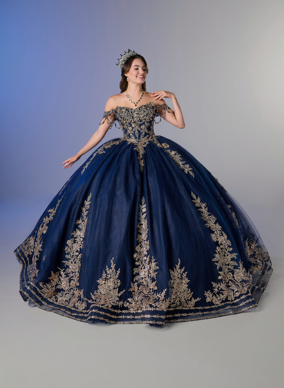 A woman modeling an elaborate off-the-shoulder ball gown in deep navy blue with extensive gold floral embroidery, set against a studio background with a blue-to-gray gradient. / Two-piece off-shoulder dress with a fitted, embellished bodice and a short, form-fitting skirt, featuring a sweetheart neckline, sparkling embroidered details, detachable arm drapes, and a full tulle skirt with a sweeping train.