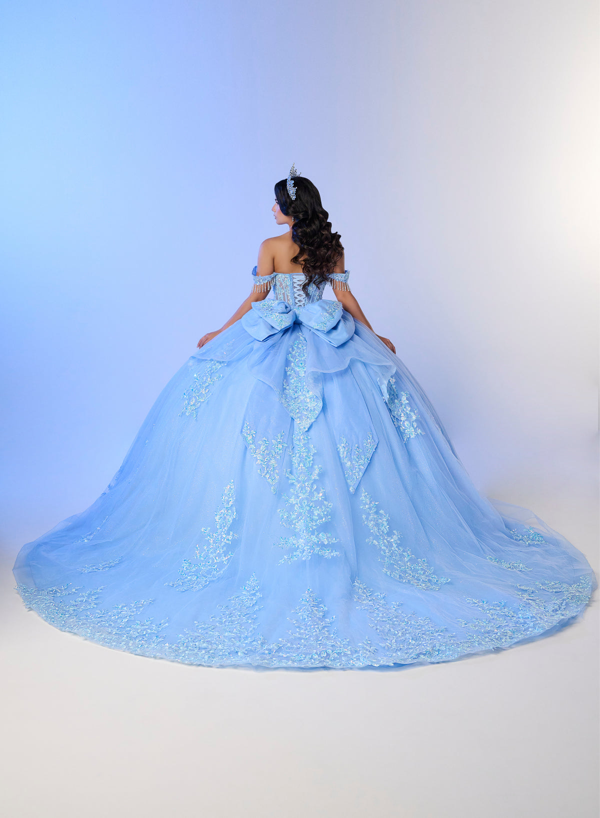 A young woman viewed from behind wearing an off-the-shoulder light blue ball gown with a very full skirt, large bow at the back, and embroidered details, posed against a soft blue-to-white studio backdrop. / Luxurious off-the-shoulder ball gown with intricate detailing and a statement bow at the back.