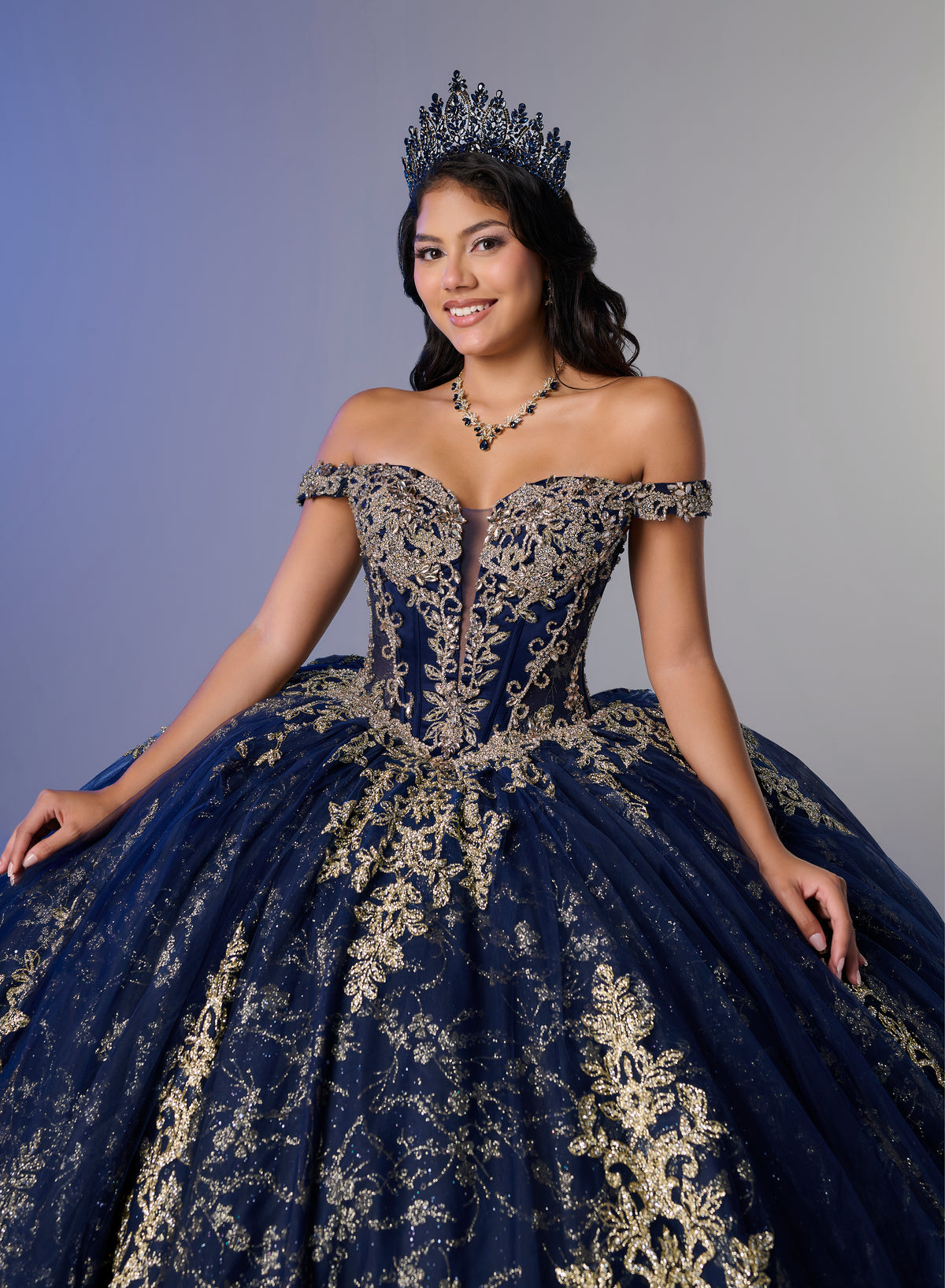 A woman wearing an elaborate off-the-shoulder navy blue ball gown with ornate gold embroidery, a matching tiara, and a necklace, posed against a plain gray studio background. / Elegant off-shoulder ball gown featuring intricate embroidered detailing, a structured bodice, and a voluminous skirt with a dramatic statement bow at the back.