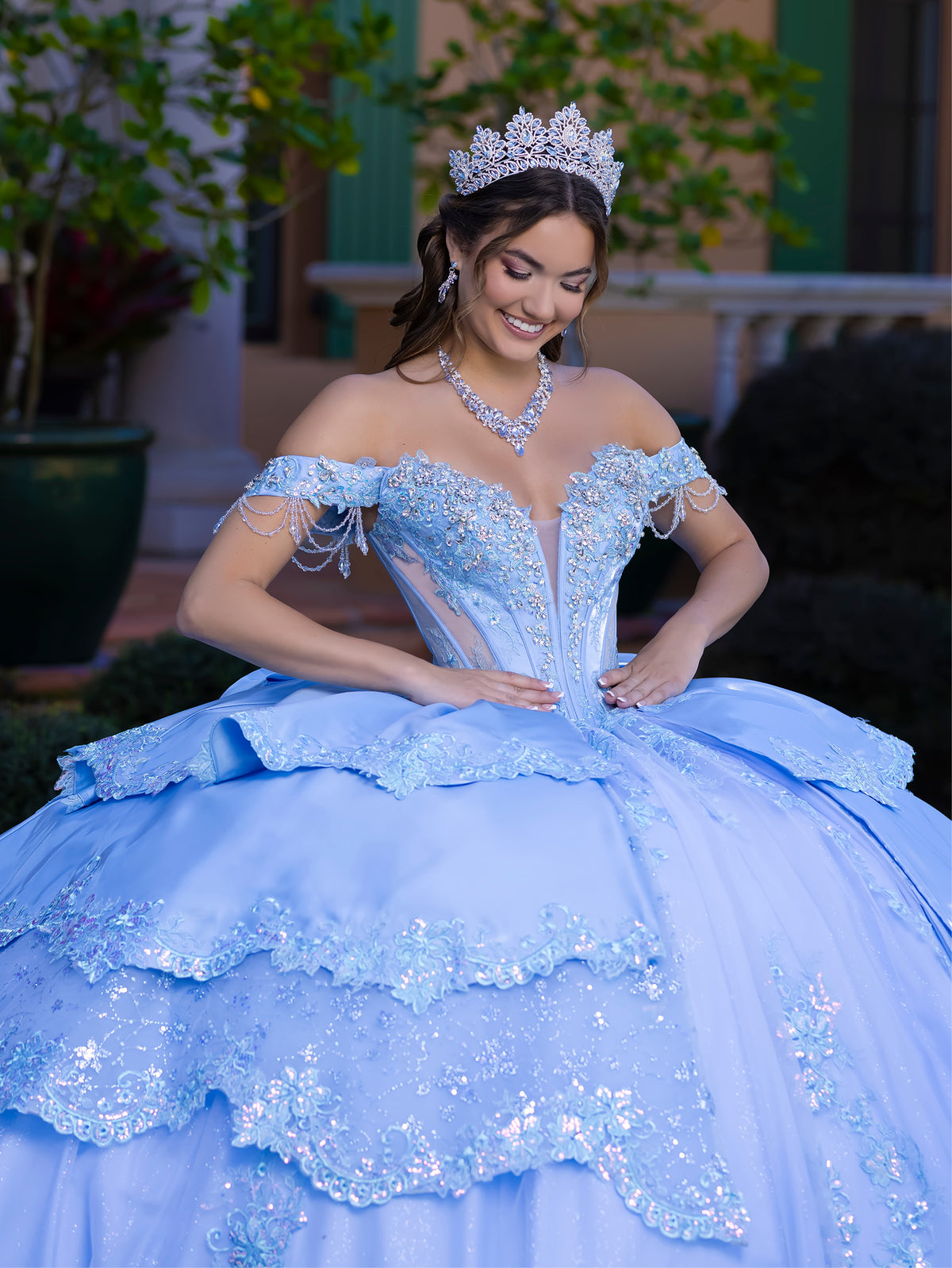Woman in a light blue quinceañera dress with a tiara, standing outdoors. / This gown features a fitted, embellished bodice with off-the-shoulder sleeves and a full tiered ball-gown skirt. Intricate appliqués decorate the layers, and a long flowing train with a large back bow completes the princess-style look.