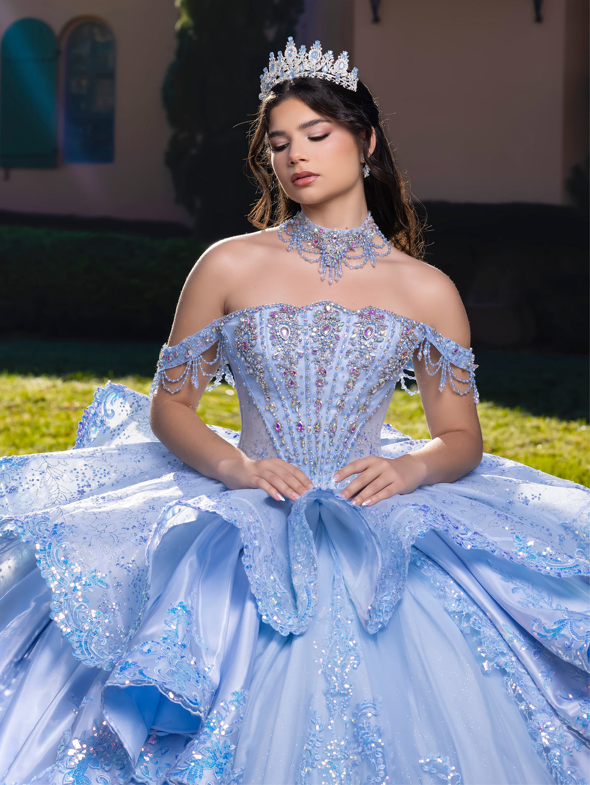 Woman in a blue sequin gown with a tiara outdoors / This gown features a detailed off-the-shoulder bodice and a full layered ball-gown skirt. A large, sculpted bow highlights the back, leading into a long train decorated with ornate appliqués for a dramatic, regal finish.