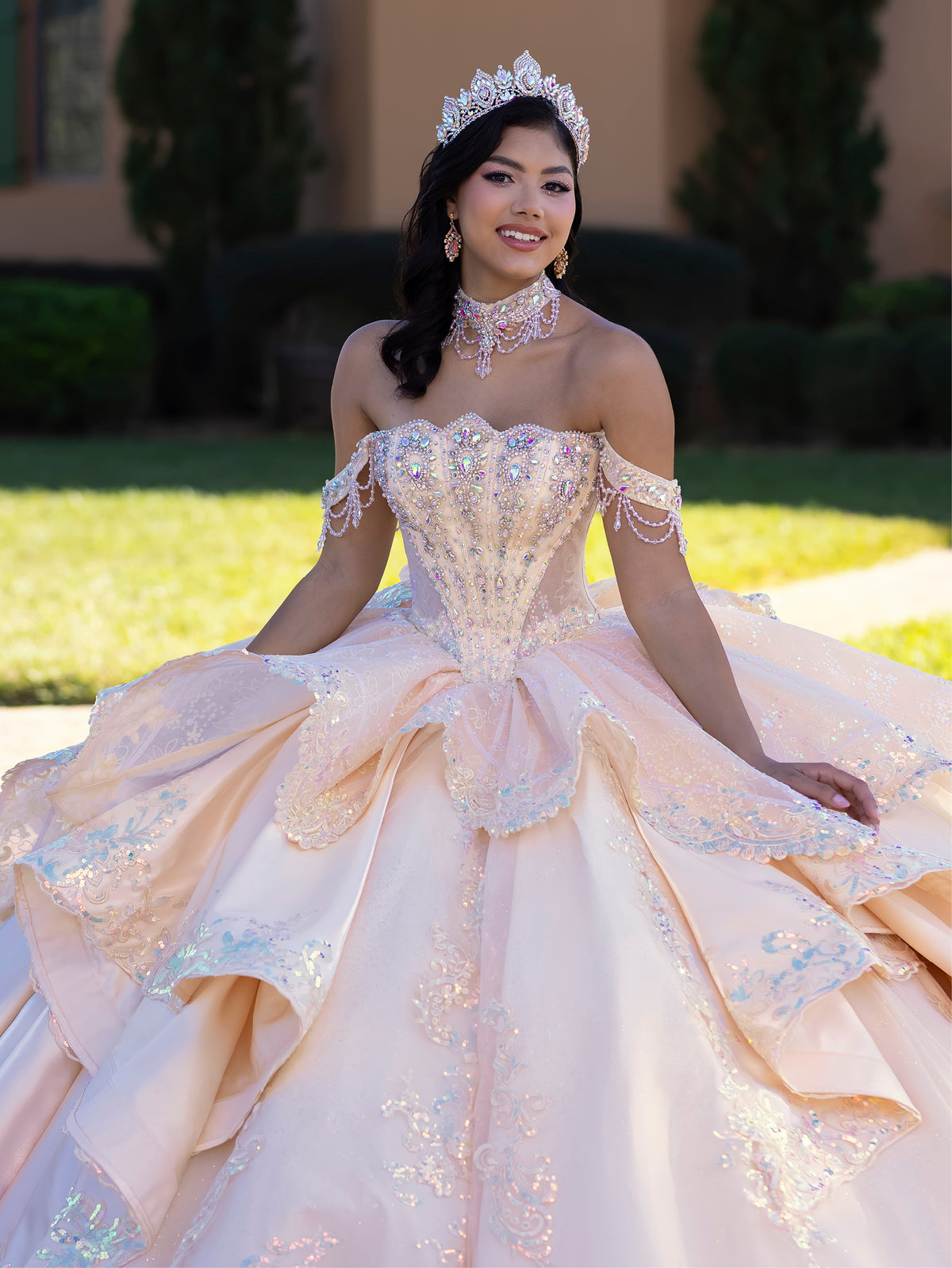 Woman in a glamorous gown with intricate details and a tiara, standing outdoors. / This gown features a detailed off-the-shoulder bodice and a full layered ball-gown skirt. A large, sculpted bow highlights the back, leading into a long train decorated with ornate appliqués for a dramatic, regal finish.