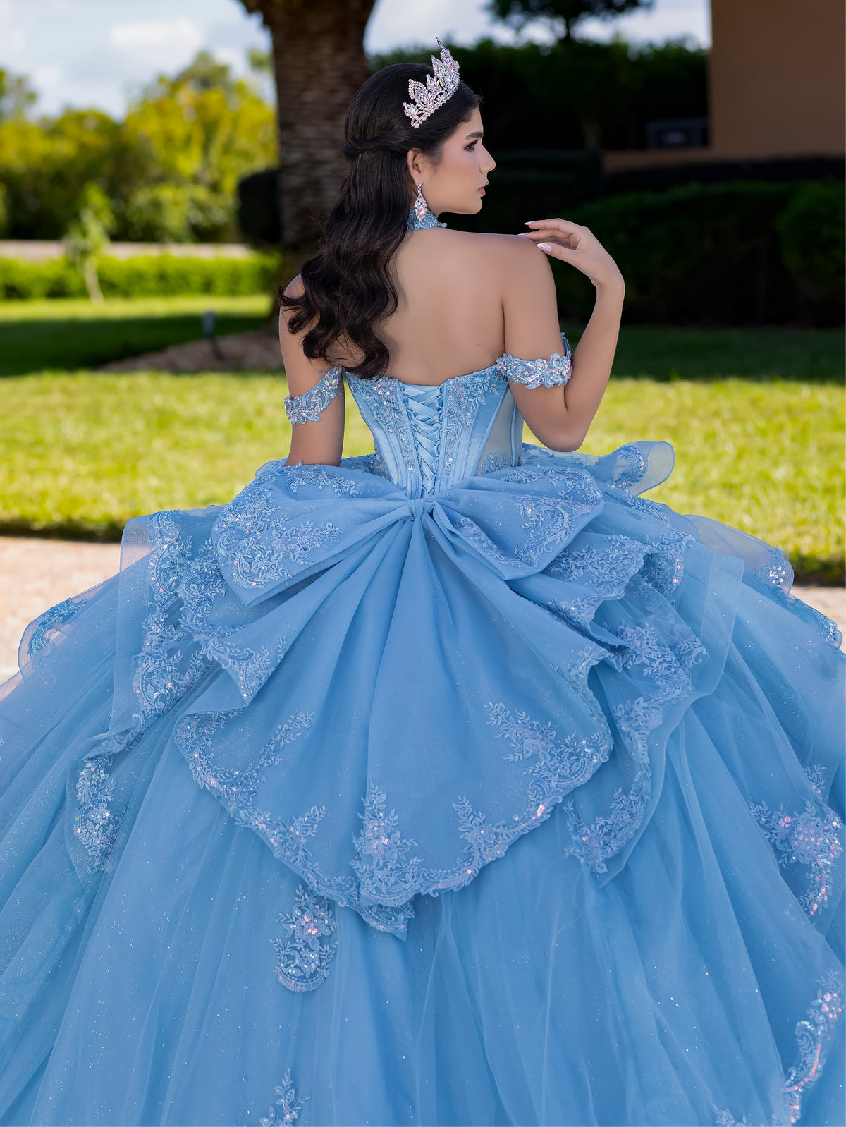 Woman in a blue quinceañera dress with intricate detailing outdoors. / This gown features a fitted, embellished bodice with off-the-shoulder straps and a full layered ball-gown skirt. Intricate appliqués decorate the skirt, while a large, sculpted bow and long ornate train complete the dramatic, regal design.