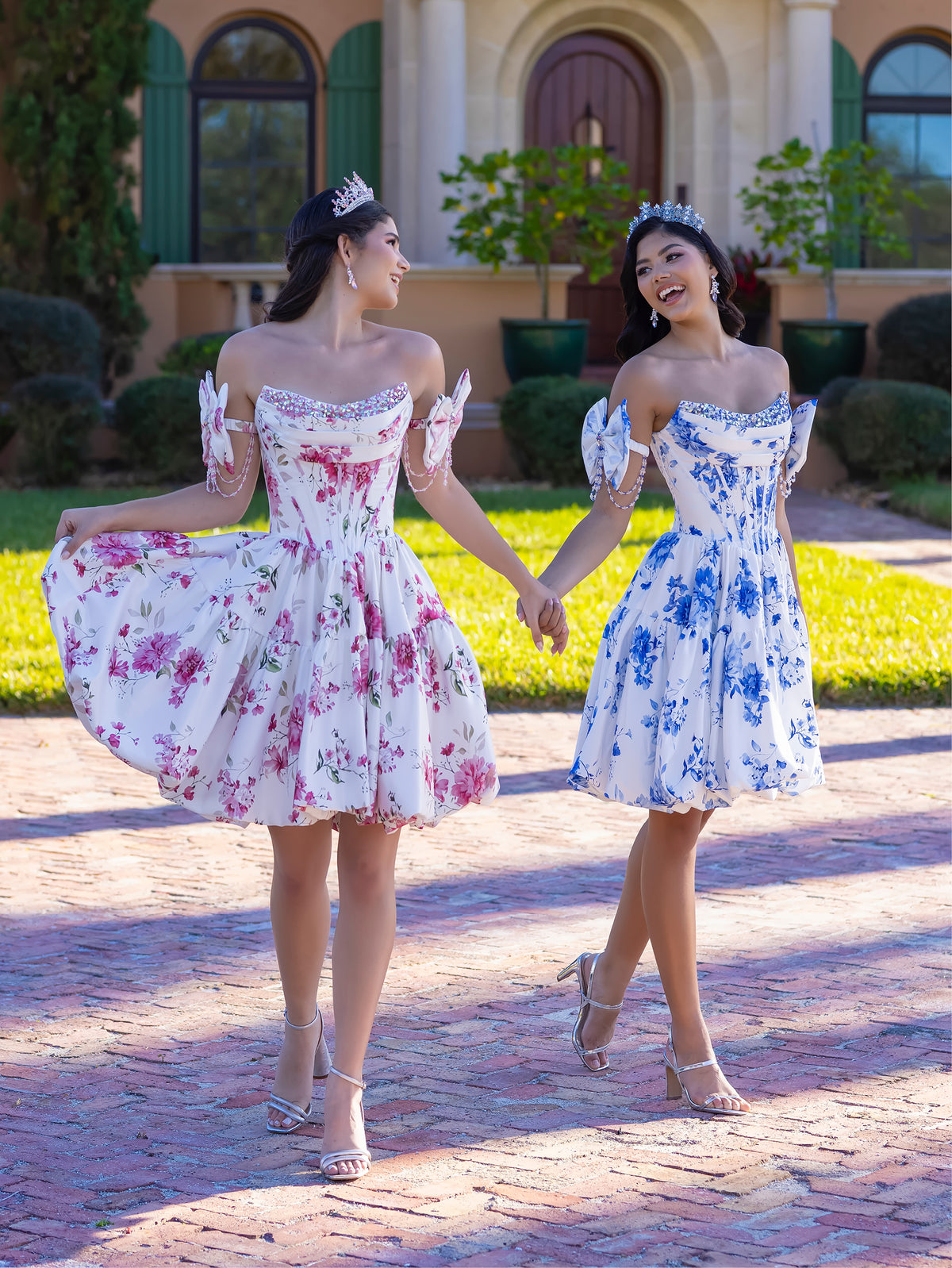 Two women in matching floral off-the-shoulder mini dresses holding hands while walking on a brick path in front of a house. One dress has a pink floral pattern on white, and the other has a blue floral pattern on white. / This gown features a full ball-gown skirt with delicate floral detailing and a fitted bodice with off-the-shoulder sleeves. A long, flowing train completes the elegant, fairytale look.