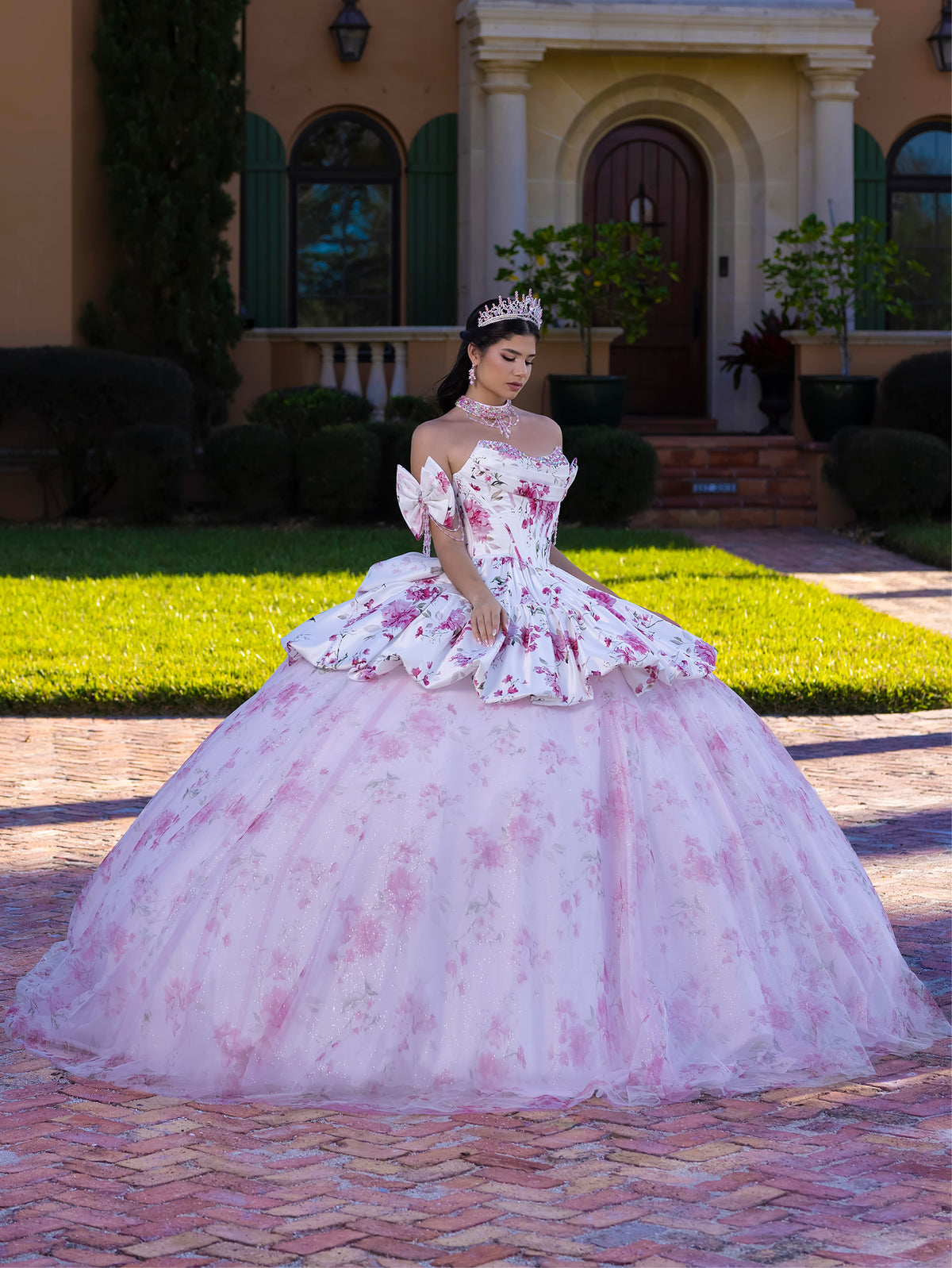 Woman in a floral quinceañera dress standing in front of a house. / This gown features a full ball-gown skirt with delicate floral detailing and a fitted bodice with off-the-shoulder sleeves. A long, flowing train completes the elegant, fairytale look.