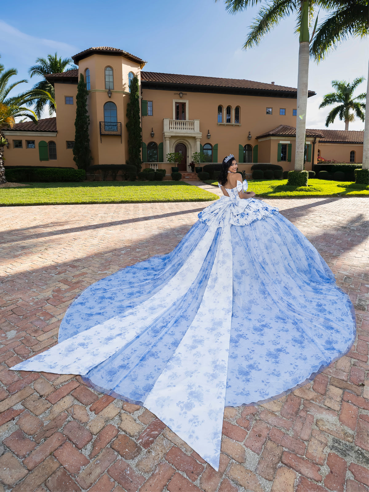 Woman in a blue gown with a long train standing in front of a large house. / This gown features a full ball-gown skirt with delicate floral detailing and a fitted bodice with off-the-shoulder sleeves. A long, flowing train completes the elegant, fairytale look.