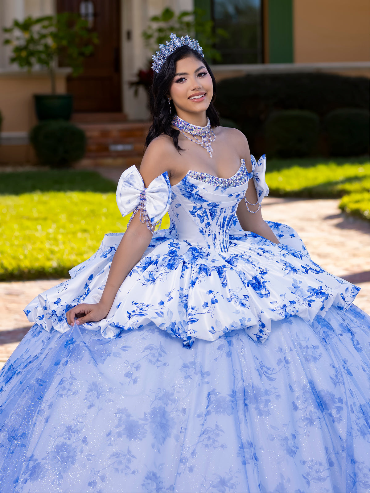 Woman in a blue and white floral dress with a tiara outdoors. / This gown features a full ball-gown skirt with delicate floral detailing and a fitted bodice with off-the-shoulder sleeves. A long, flowing train completes the elegant, fairytale look.