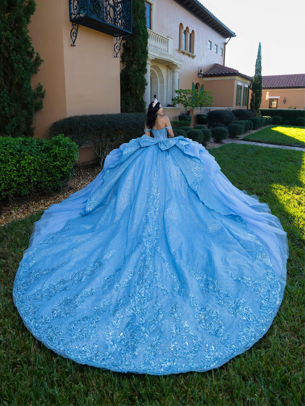 A woman photographed from behind wearing an elaborate light blue ball gown with a very large train, standing on a lawn in front of a large house. / This gown features an embellished off-the-shoulder bodice with layered textures and a full ball-gown skirt. The cascading layers create dramatic volume, while the back extends into a sweeping, embroidered train for an elegant, regal finish.