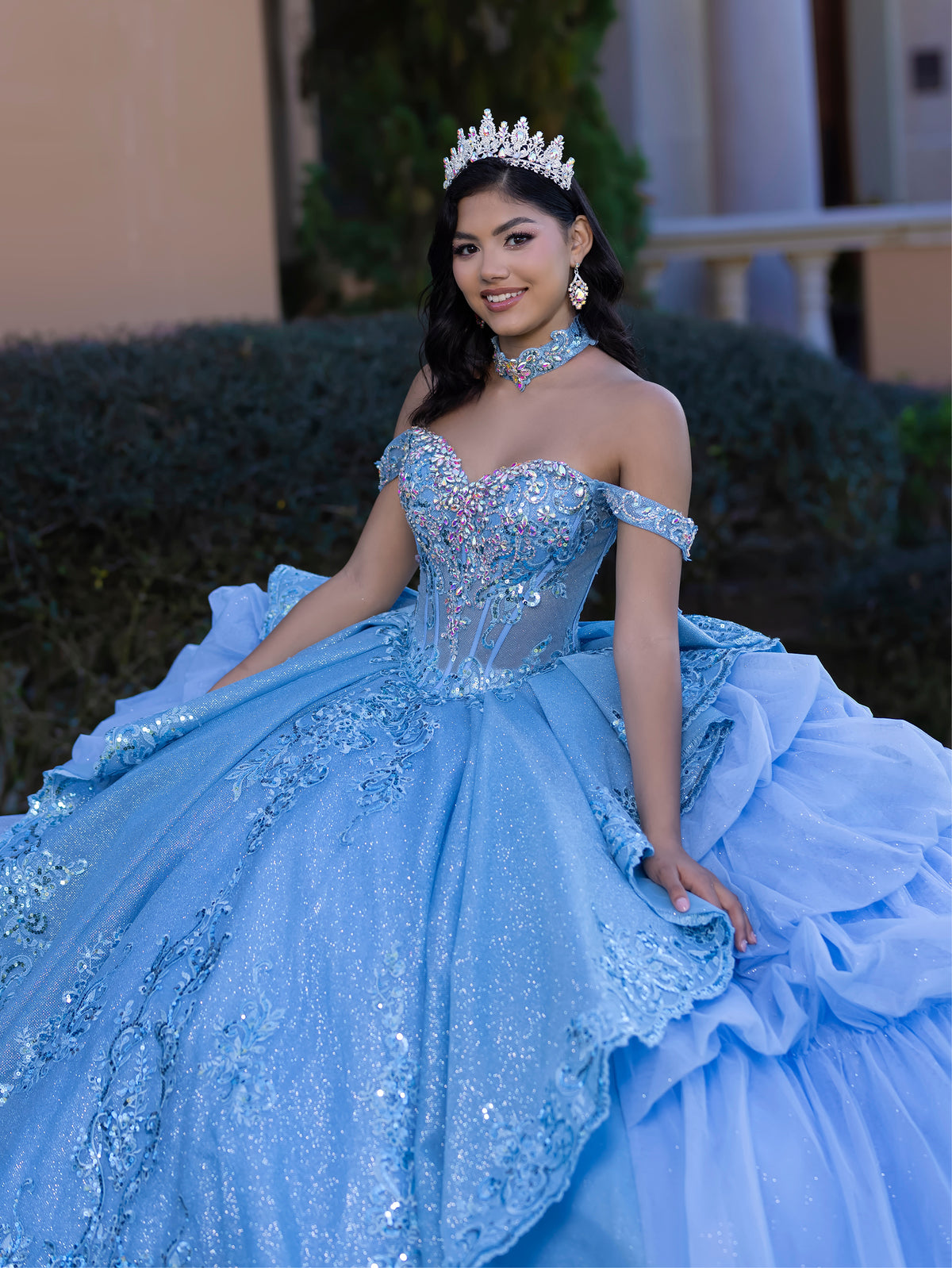 Woman in a blue quinceañera dress with a tiara and necklace, sitting outdoors. / This gown features an embellished off-the-shoulder bodice with layered textures and a full ball-gown skirt. The cascading layers create dramatic volume, while the back extends into a sweeping, embroidered train for an elegant, regal finish.