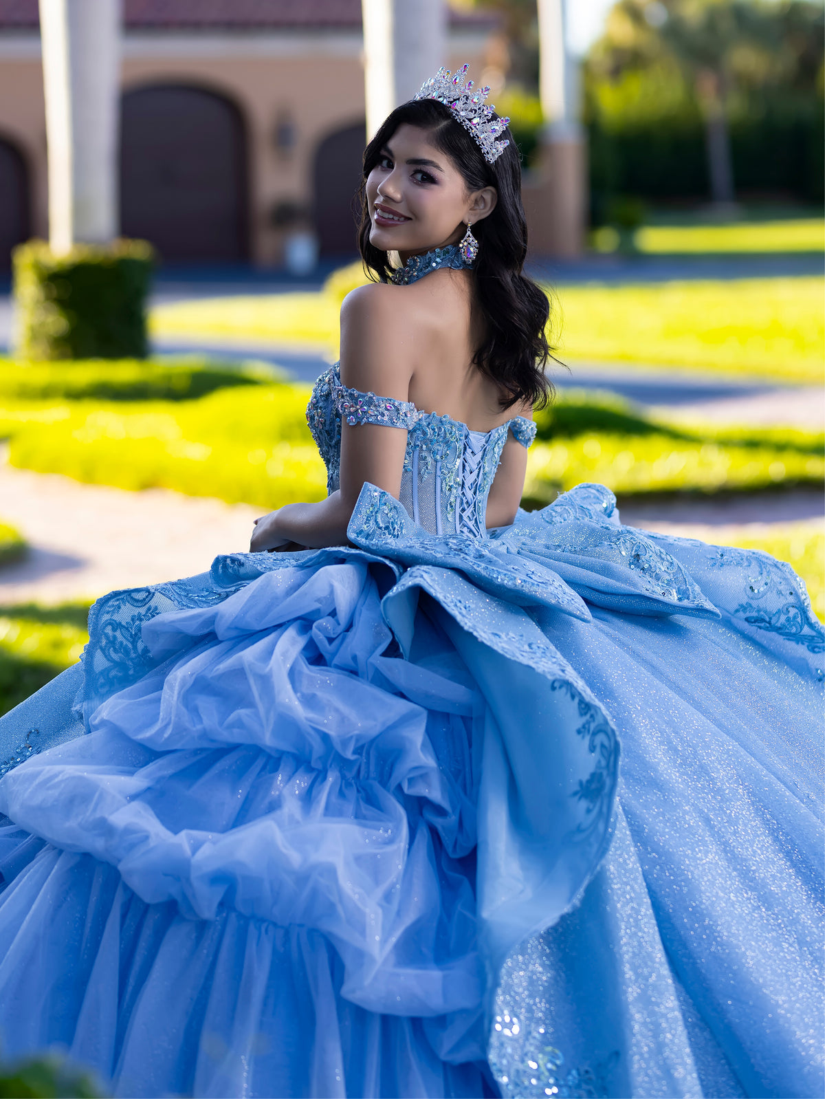 Woman in a blue formal gown with intricate details outdoors. / This gown features an embellished off-the-shoulder bodice with layered textures and a full ball-gown skirt. The cascading layers create dramatic volume, while the back extends into a sweeping, embroidered train for an elegant, regal finish.