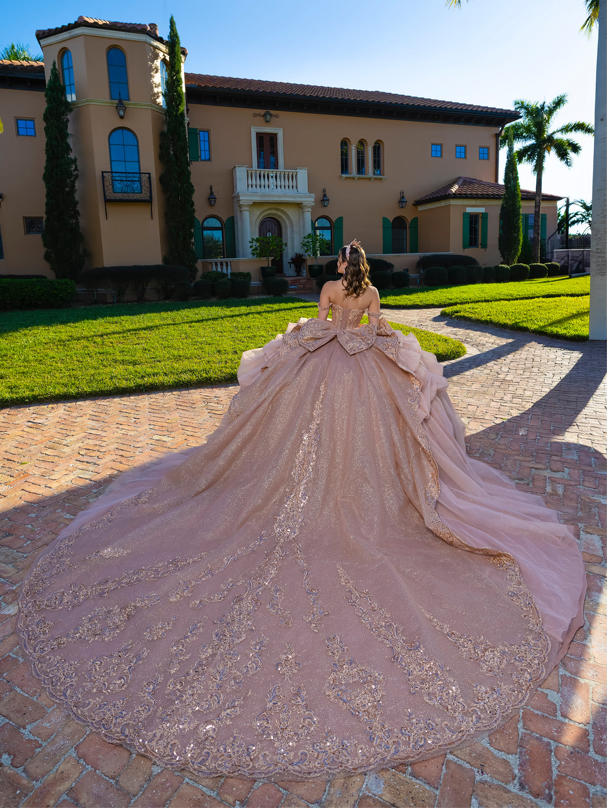A woman stands outdoors in front of a large beige mansion on a stone driveway, wearing an elaborate ball gown with a very long train. The dress is a dusty blush pink with intricate gold beading and embroidery. / This gown features an embellished off-the-shoulder bodice with layered textures and a full ball-gown skirt. The cascading layers create dramatic volume, while the back extends into a sweeping, embroidered train for an elegant, regal finish.