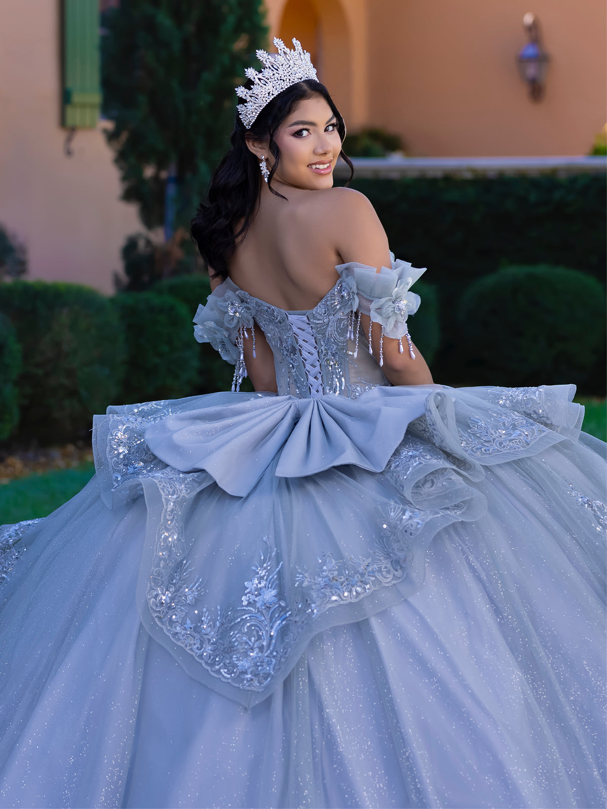 Woman in a sparkling blue gown with a large bow, wearing a tiara, outdoors. / This gown features an embellished off-the-shoulder bodice with delicate appliqué and a full tulle skirt decorated with floral embroidery. A structured accent at the waist and a long, ornate train complete the dramatic look.