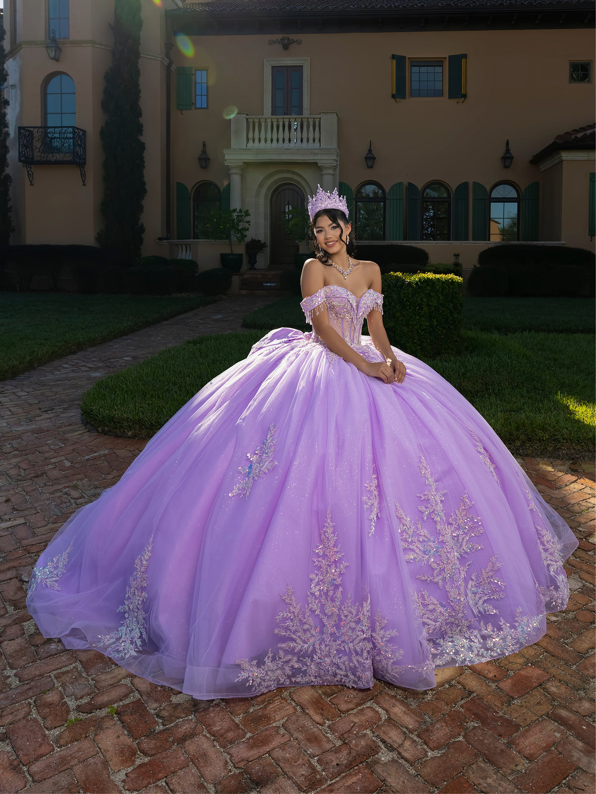 A woman wearing a voluminous lavender off-the-shoulder ball gown with silver floral embroidery and a matching tiara, standing on a brick path in front of a large villa. / This ball gown features an embellished off-the-shoulder bodice, and a full skirt decorated with intricate floral appliqué, finished with a back bow.