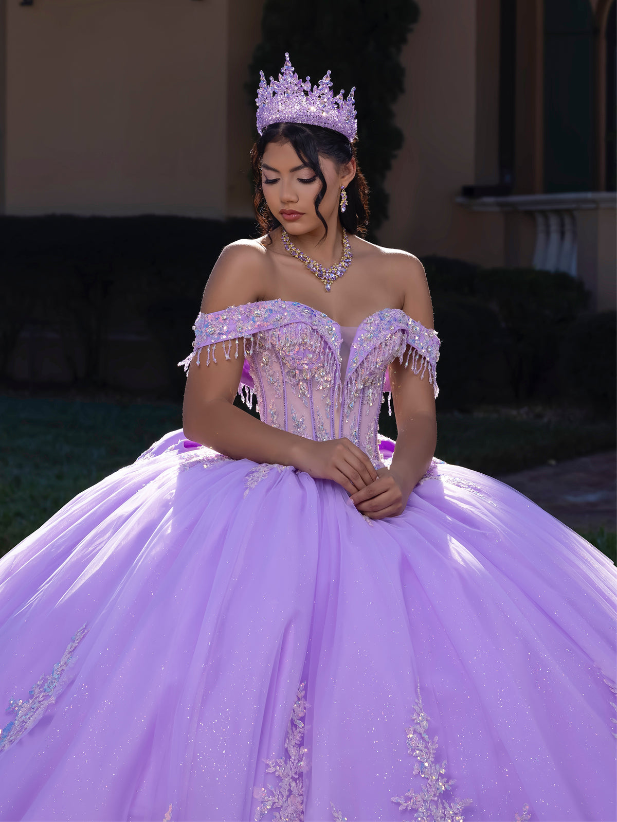 Woman in a purple quinceañera dress with a tiara, standing outdoors. / This ball gown features an embellished off-the-shoulder bodice, and a full skirt decorated with intricate floral appliqué, finished with a back bow.