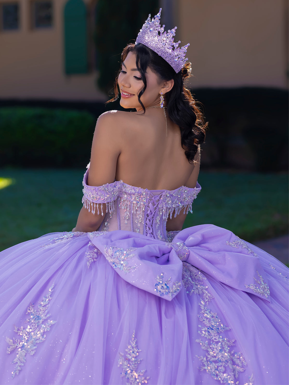 A woman wearing a lavender off-the-shoulder quinceañera ball gown with a large bow at the back, embellished with sparkling silver and blue beading, and wearing a matching lavender tiara, photographed outdoors. / This ball gown features an embellished off-the-shoulder bodice, and a full skirt decorated with intricate floral appliqué, finished with a back bow.