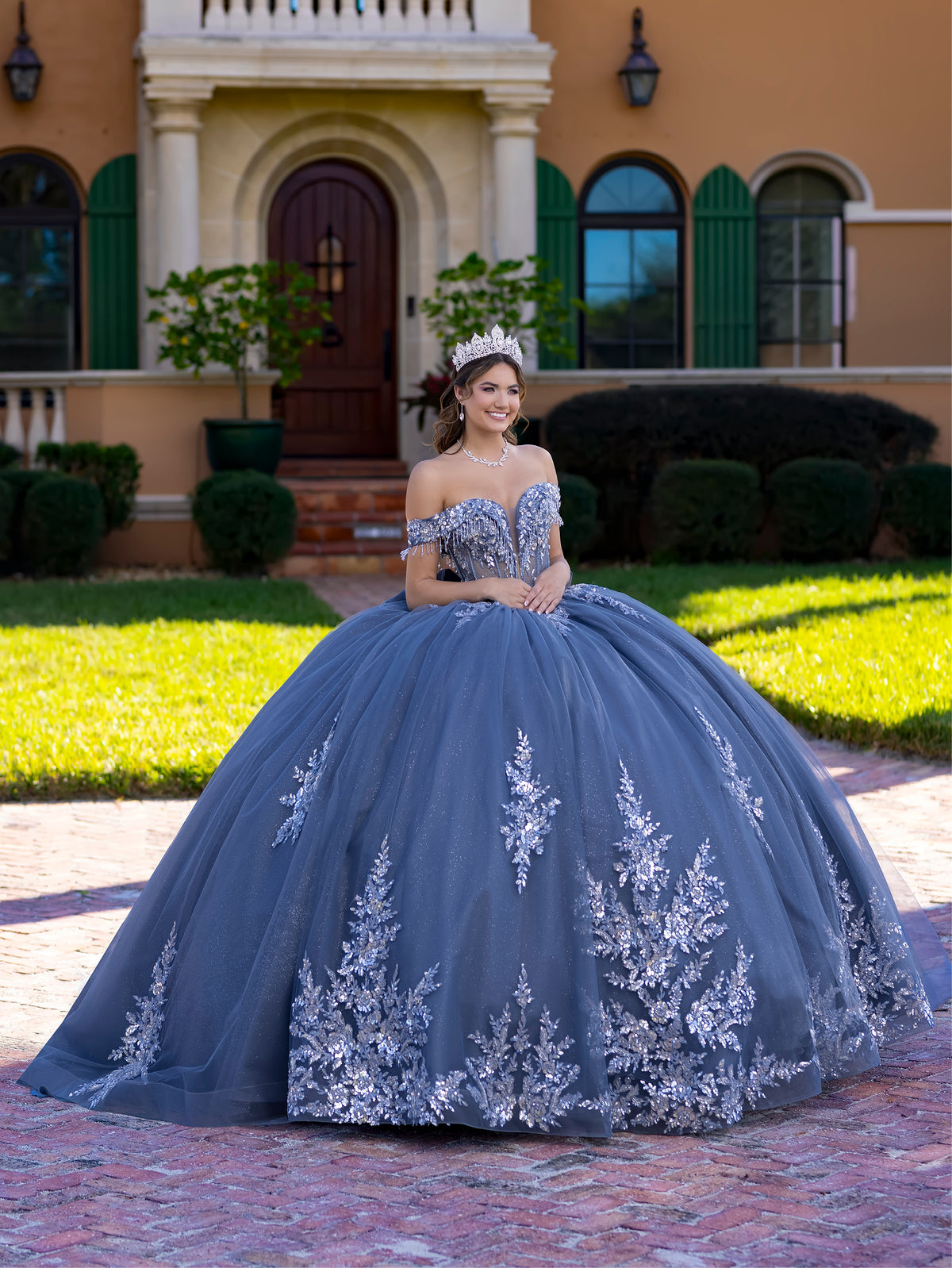Woman in a blue quinceañera dress with floral embroidery standing in front of a building. / This ball gown features an embellished off-the-shoulder bodice, and a full skirt decorated with intricate floral appliqué, finished with a back bow.