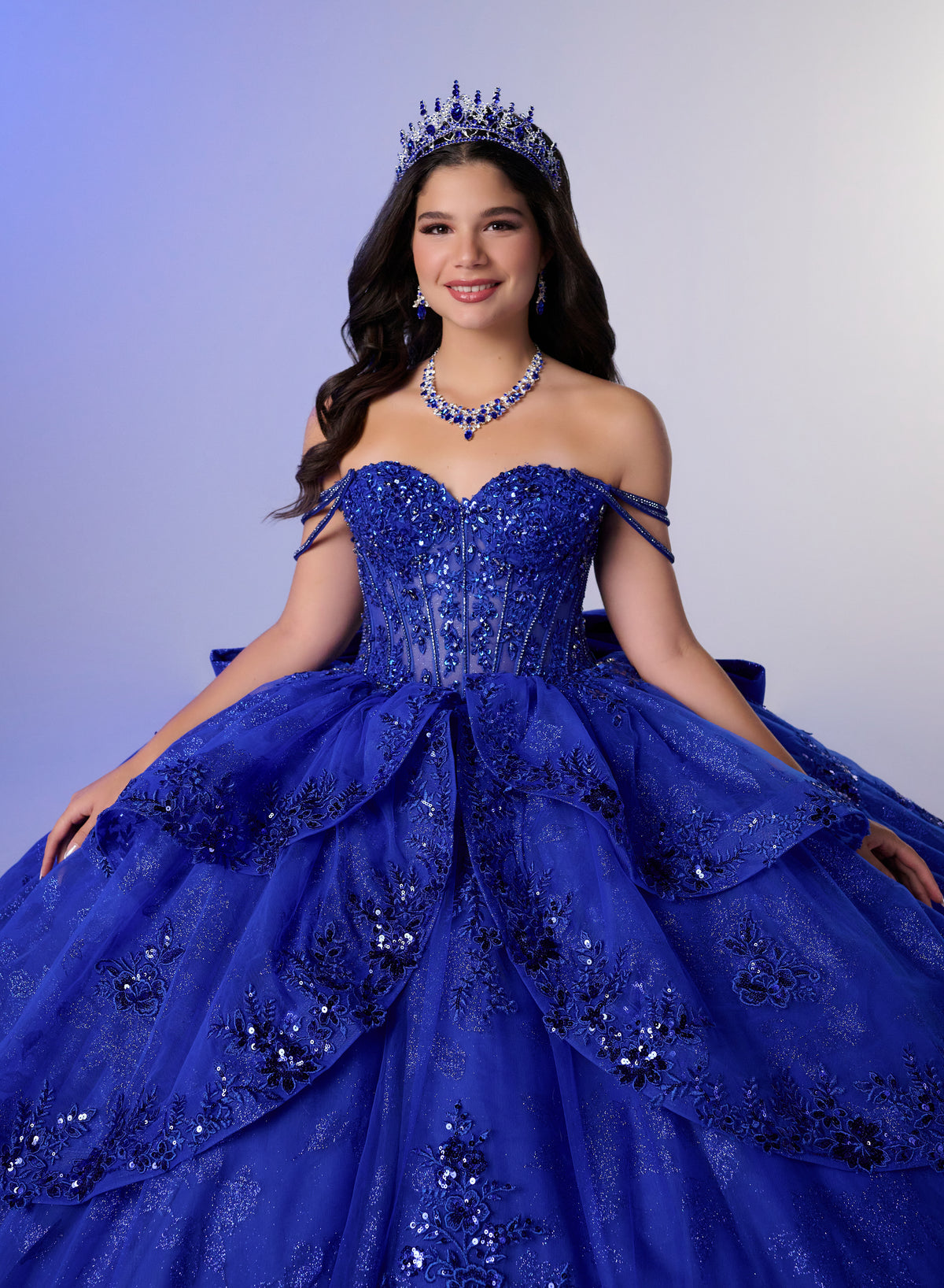 A young woman wearing an elaborate royal blue ball gown with beaded detailing, a matching tiara, and a blue beaded necklace, posed against a soft blue-to-gray gradient studio background. / Glamorous ball gown featuring a sparkling embellished corset bodice, dramatic layered overskirt details, a voluminous skirt, and an elegant sweeping train.