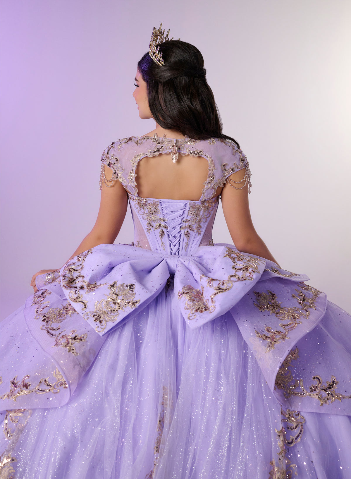 A woman shown from the back wearing an elaborate lavender ball gown with gold embroidery, a large bow at the back, sheer sleeves, and a jeweled crown against a soft lavender studio background. / Regal ball gown featuring intricate embroidered appliqués, a structured bodice, dramatic layered overskirt details, and a sweeping cathedral-length train for a royal-inspired look.
