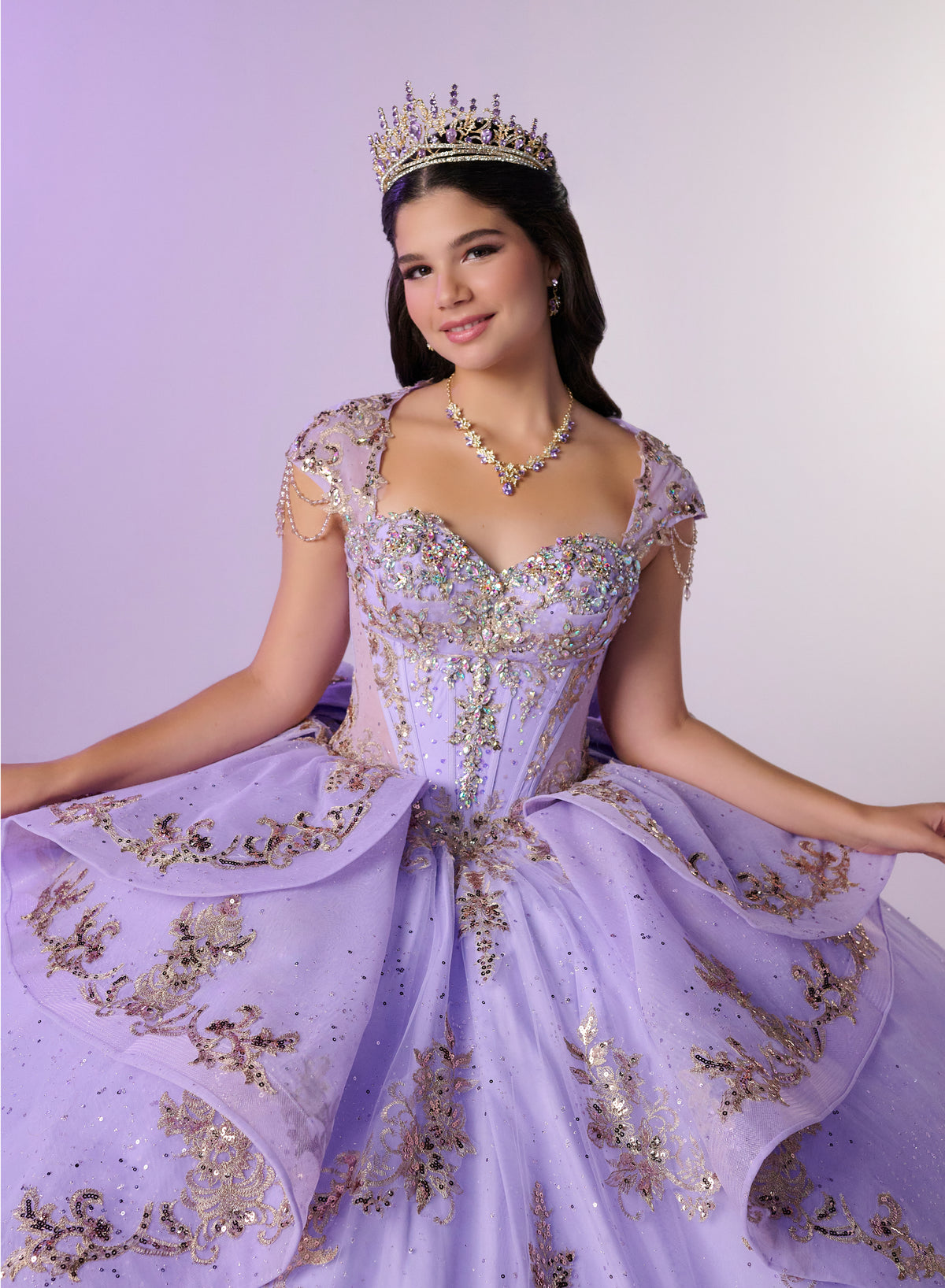 A young woman wearing an elaborate lavender quinceañera dress with gold sequin embroidery, a matching tiara, and a necklace, posing against a light purple studio background. / Regal ball gown featuring intricate embroidered appliqués, a structured bodice, dramatic layered overskirt details, and a sweeping cathedral-length train for a royal-inspired look.