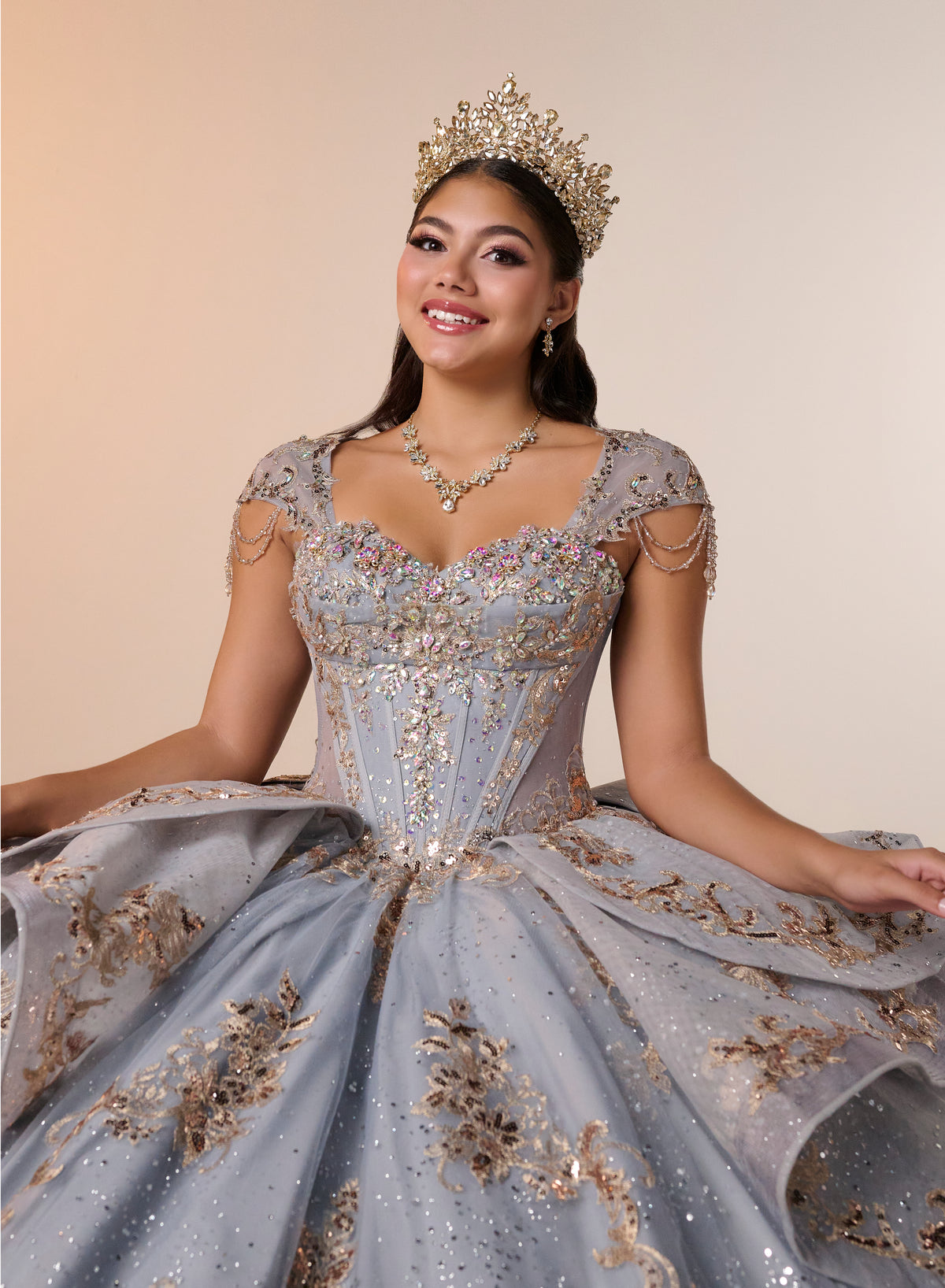A woman wearing an ornate light blue ball gown with gold embroidery, a matching tiara, and a necklace, posing against a solid beige background. / Regal ball gown featuring intricate embroidered appliqués, a structured bodice, dramatic layered overskirt details, and a sweeping cathedral-length train for a royal-inspired look.