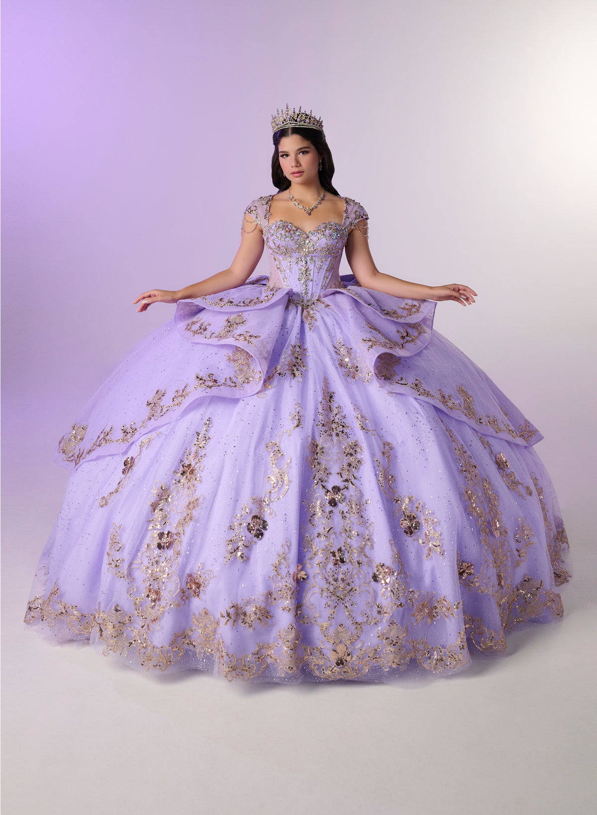 A woman wearing a voluminous lavender quinceañera ball gown with gold floral embroidery and a tiara, posed against a light lavender studio backdrop. / Regal ball gown featuring intricate embroidered appliqués, a structured bodice, dramatic layered overskirt details, and a sweeping cathedral-length train for a royal-inspired look.