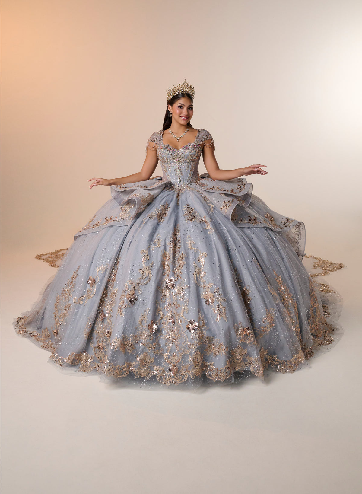 A woman wearing a voluminous light blue quinceañera ball gown with gold floral embroidery and a matching tiara, posed against a warm beige studio backdrop. / Regal ball gown featuring intricate embroidered appliqués, a structured bodice, dramatic layered overskirt details, and a sweeping cathedral-length train for a royal-inspired look.