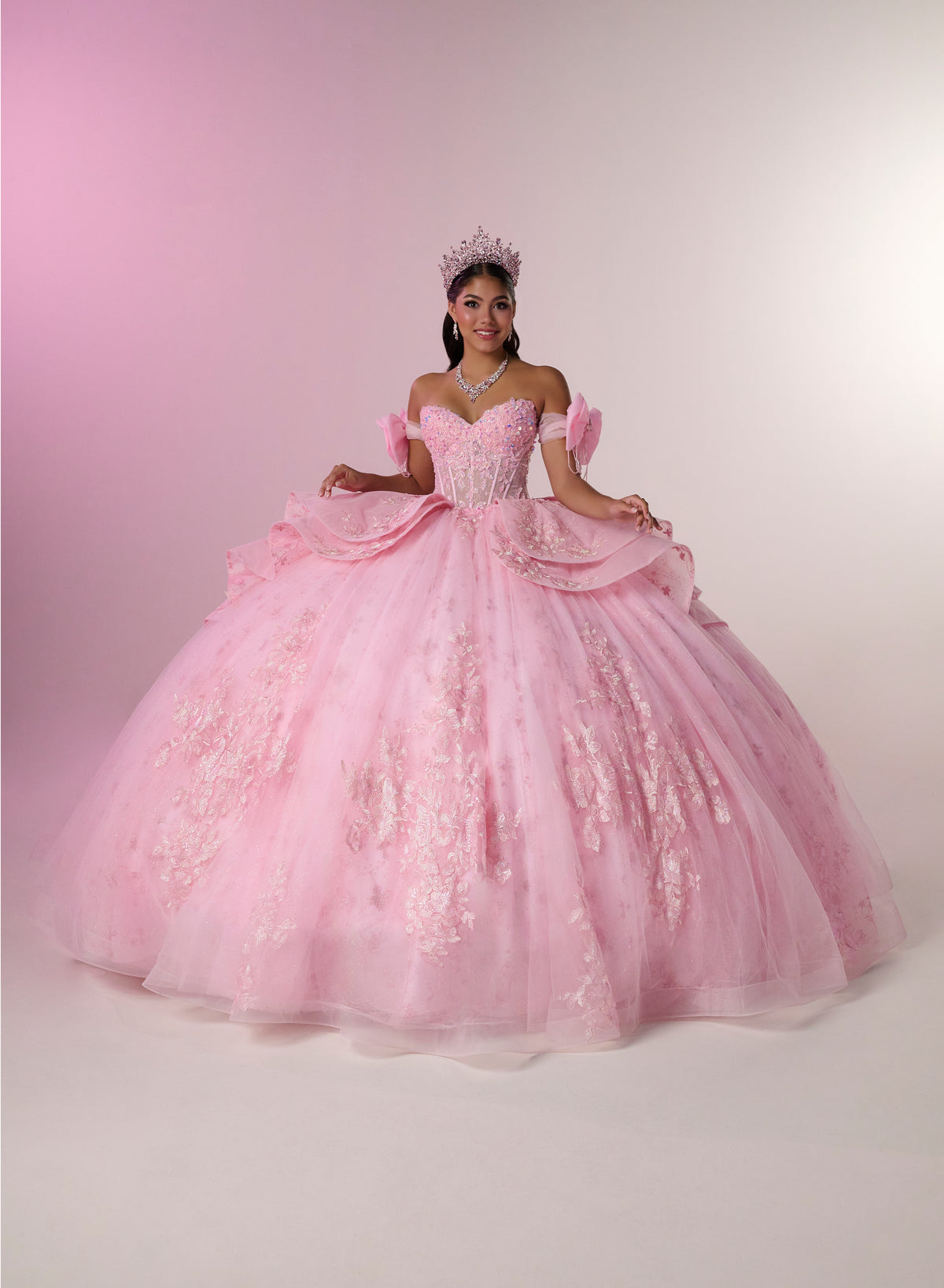 A woman wearing a voluminous light pink ball gown with off-the-shoulder puff sleeves, a fitted embellished bodice, and a full layered tulle skirt, posed against a soft pink-to-white studio backdrop. She is also wearing a tiara. / Princess-inspired ball gown featuring a sweetheart corset bodice, intricate floral appliqués, dramatic layered overskirt details, and a voluminous skirt with a statement bow at the back.