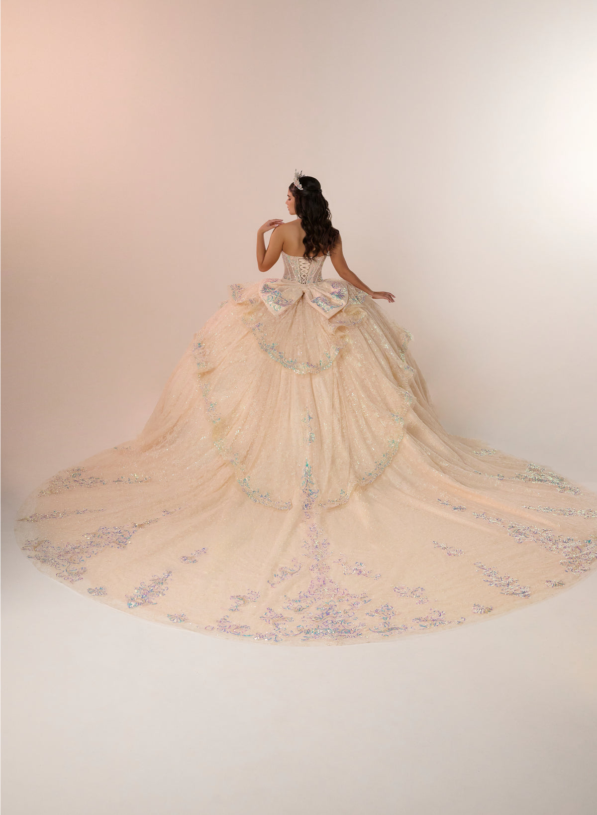 A woman stands with her back to the camera wearing an elaborate off-white ball gown with a very long train, decorated with pastel blue and lavender floral embroidery and beadwork, set against a plain light beige studio background. / Enchanting ball gown featuring a structured sweetheart corset bodice, shimmering embroidered details, dramatic layered ruffles, and a sweeping cathedral-length train for a regal princess-inspired look.