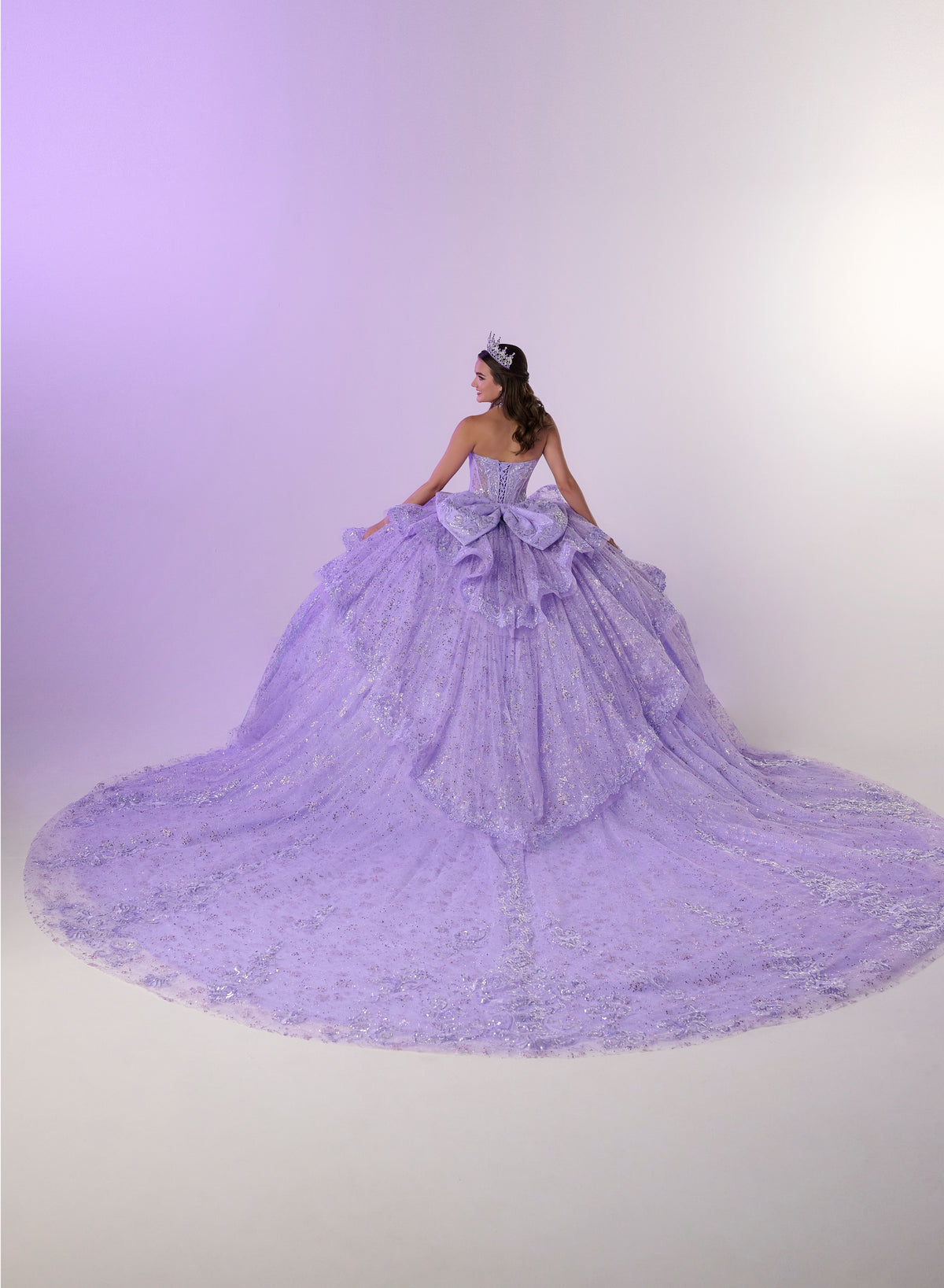 A woman wearing a voluminous lavender quinceañera dress with a long train, photographed against a soft purple studio backdrop. / Enchanting ball gown featuring a structured sweetheart corset bodice, shimmering embroidered details, dramatic layered ruffles, and a sweeping cathedral-length train for a regal princess-inspired look.