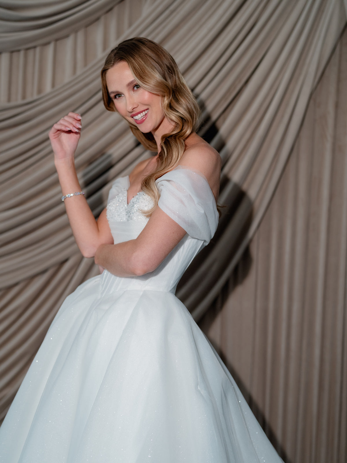 A woman wearing an off-the-shoulder white wedding gown with a voluminous sparkle tulle skirt, posing in front of draped beige fabric. / Ball gown in two face organza with glitzy underlayer, sweetheart neckline and off shoulder straps. Lace up back closure.