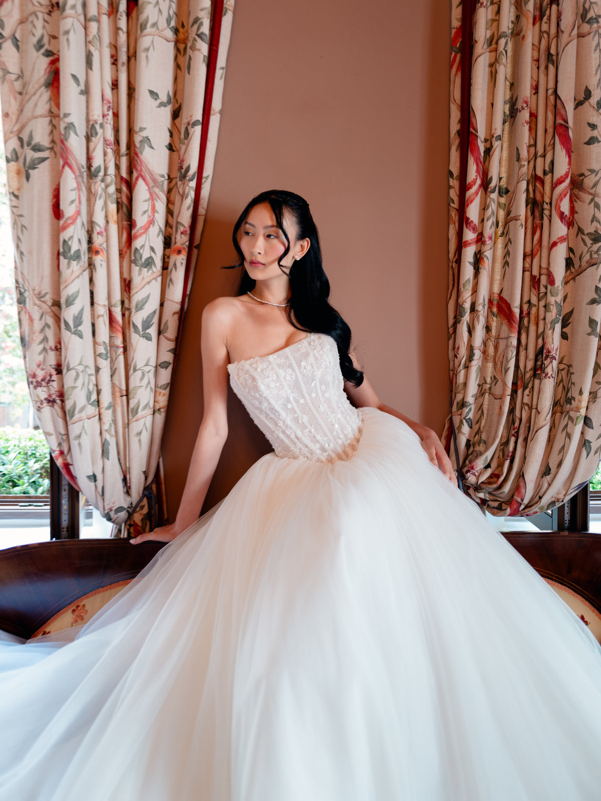 A woman wearing a strapless white wedding gown with a structured lace bodice and voluminous tulle skirt, posed indoors in front of patterned curtains and a brown wall. / Semi sheer basque waist bodice with lace appliques and dramatic tulle skirt.