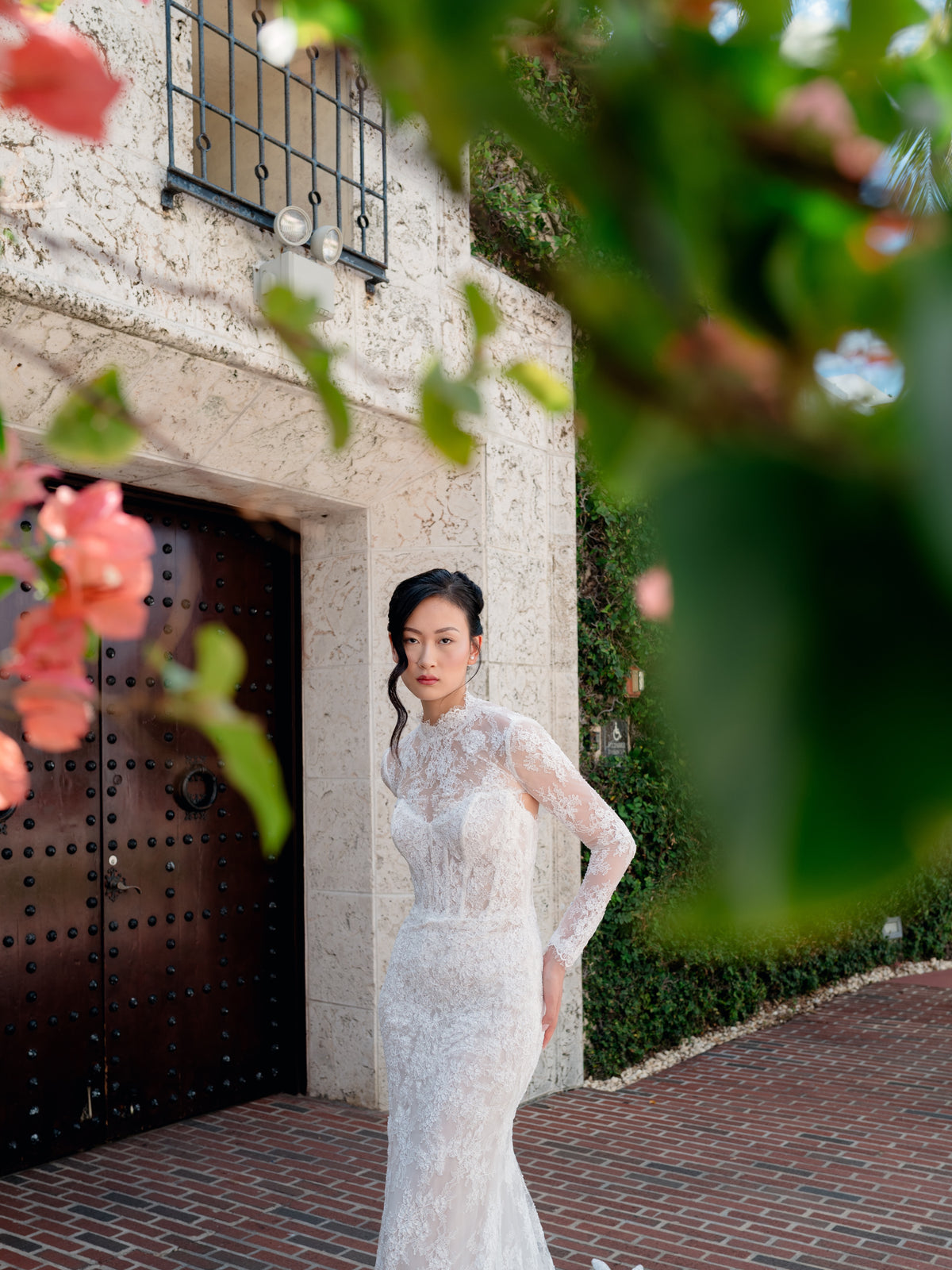 A woman wearing a fitted white lace wedding gown stands outdoors in front of a stucco building with a dark wooden door, framed by green leaves and pink flowers in the foreground. / All over lace and lace applique slim gown with detachable lining and jacket as well as structured corset.