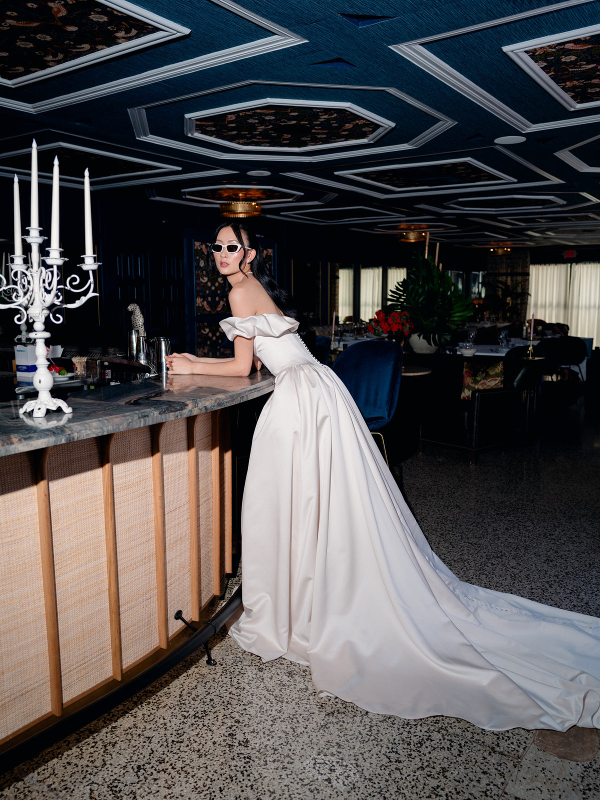 A woman in a dramatic off-the-shoulder white satin gown with a long train leans against a bar counter in an elegant, dimly lit venue with a dark patterned ceiling and a white candelabra in the foreground. / Off the shoulder sculpted neckline gown with basque waist and full, gathered skirt.